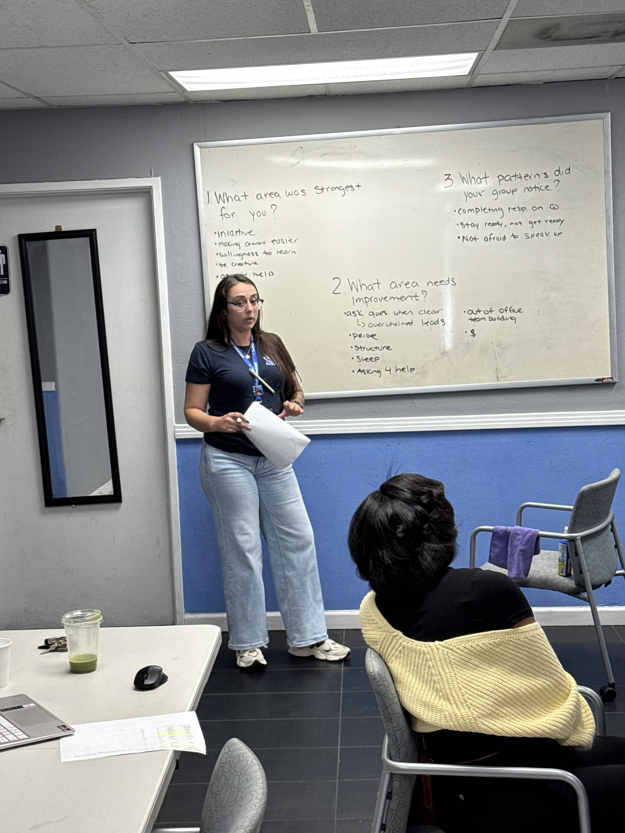A woman is standing in front of a whiteboard speaking in a classroom or meeting room, with a group of people seated and listening. The whiteboard has handwritten notes or questions. There is a table with a laptop, a mouse, some papers, and a drink in the foreground.