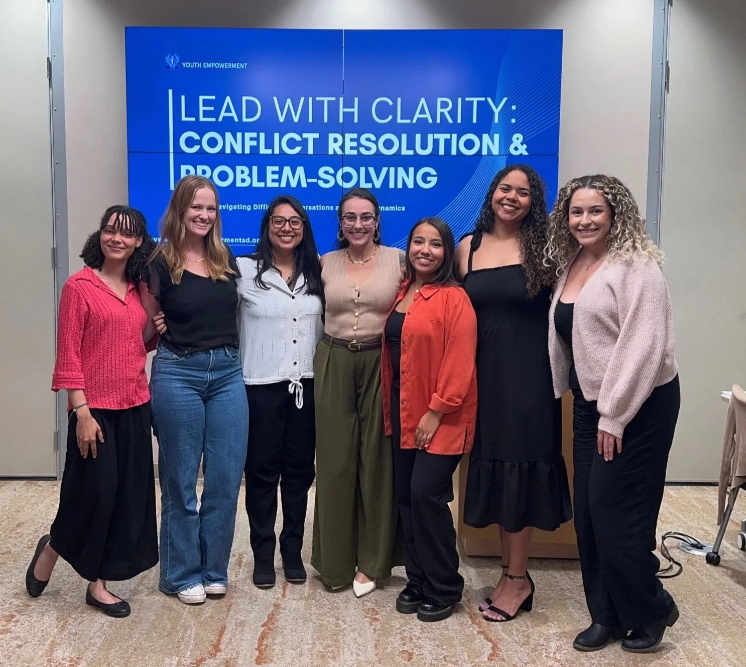 Group of seven women standing together in front of a blue presentation screen that reads 'Lead with Clarity: Conflict Resolution & Problem-Solving'.