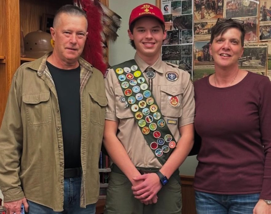 Three people standing indoors, two adults and a teenage boy in a Boy Scouts uniform with badges, smiling at the camera.