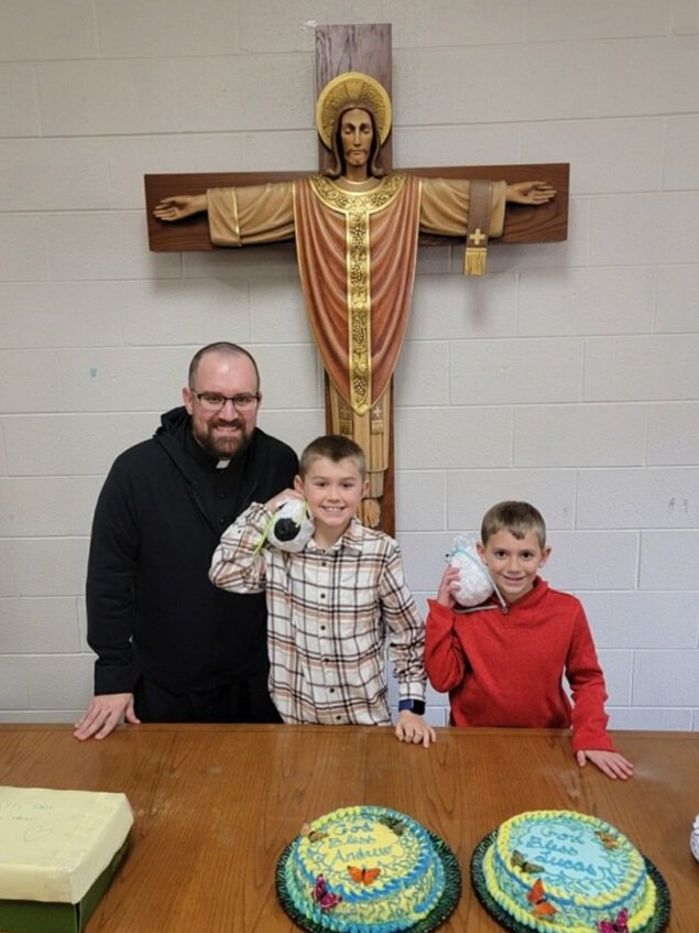 A priest and two boys stand at a table with two birthday cakes, in front of a large crucifix of Jesus on the wall.
