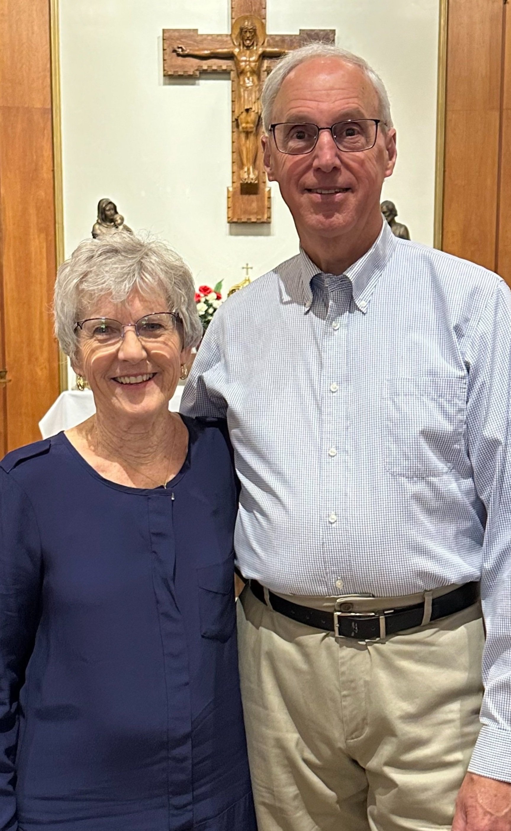 An elderly woman and man standing together inside a church, smiling at the camera. The woman wears a blue dress and glasses, and the man wears a light-colored button-up shirt and khakis. There is a crucifix and religious statues on the wall behind th