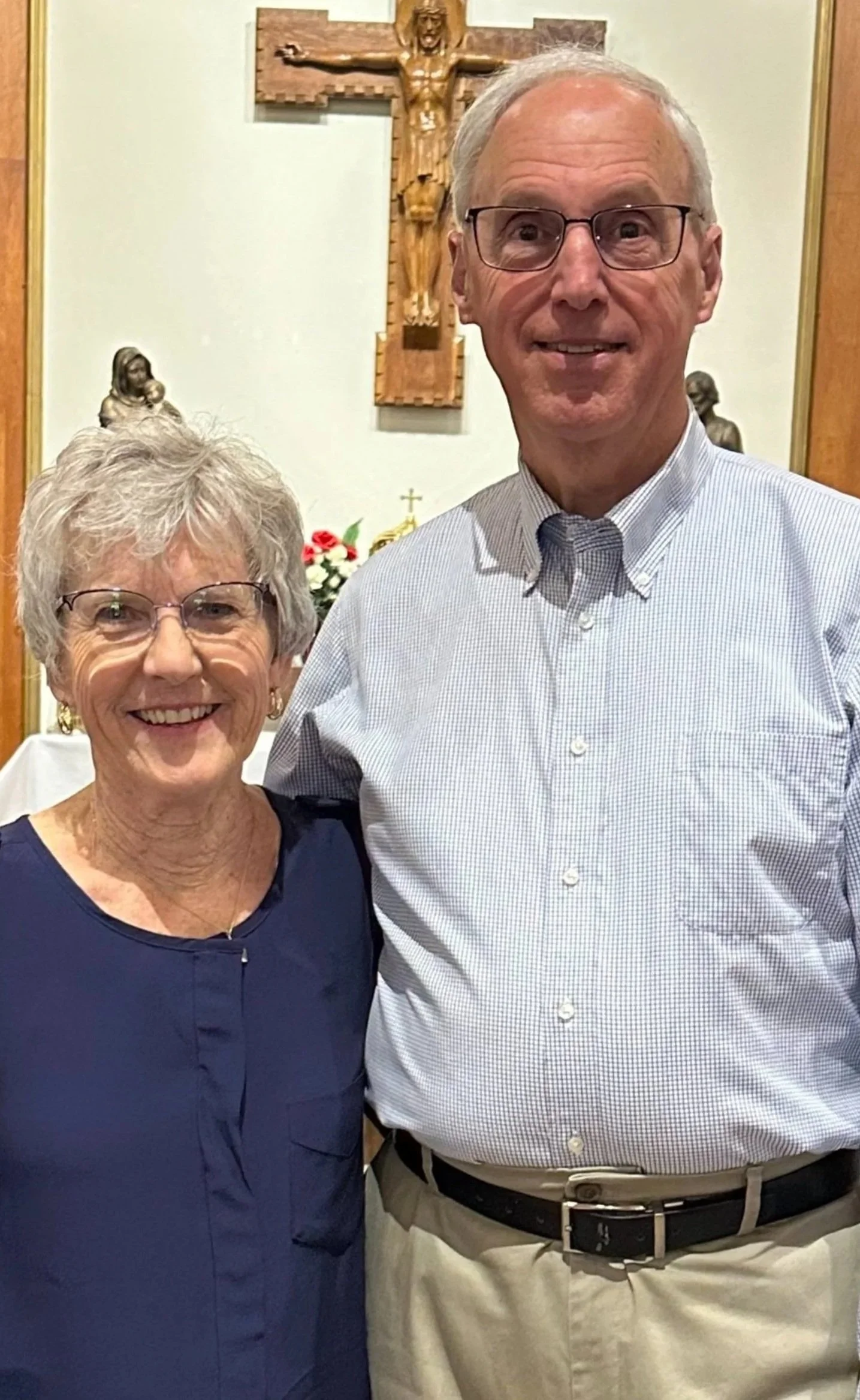 An elderly woman and man standing together inside a church, smiling at the camera. The woman wears a blue dress and glasses, and the man wears a light-colored button-up shirt and khakis. There is a crucifix and religious statues on the wall behind th
