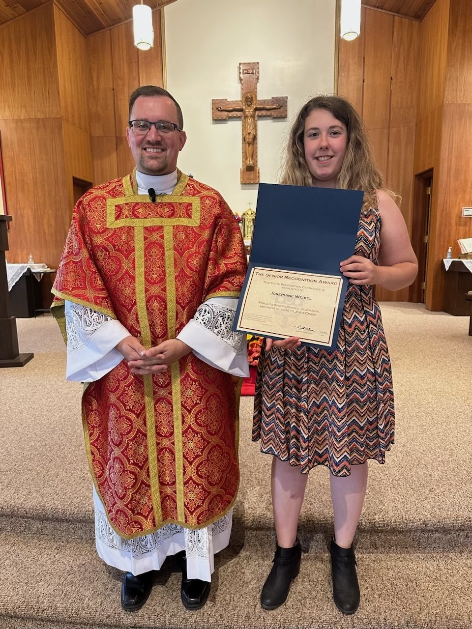 A priest in ornate red and gold liturgical vestments stands with a young girl in a patterned dress and black shoes. The girl is holding a large certificate and a folder, smiling at the camera inside a church with a wooden cross on the wall behind the