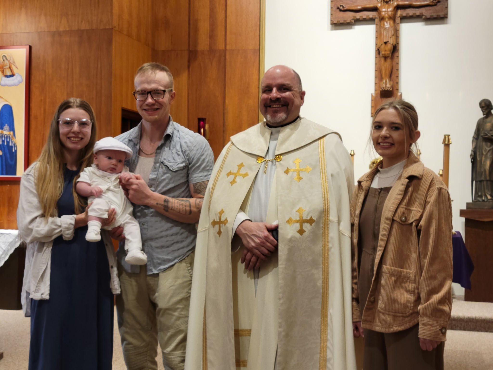 Group of people in a church, including a priest in white religious robes with gold accents, a woman with glasses holding a baby, a man with glasses, and a young woman in casual clothing, all smiling.