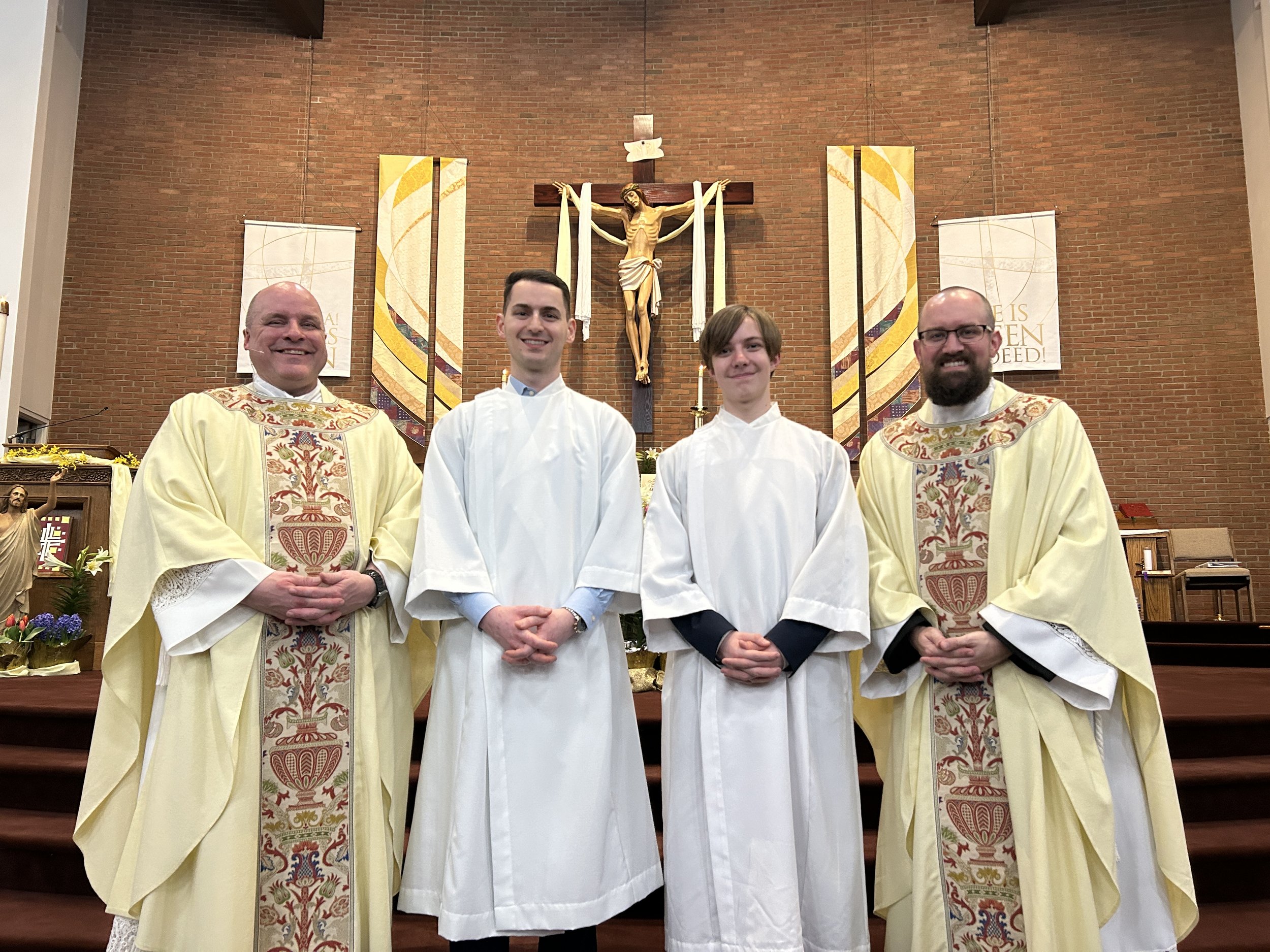 Two priests and two altar servers standing in front of a crucifix inside a church, smiling at the camera.