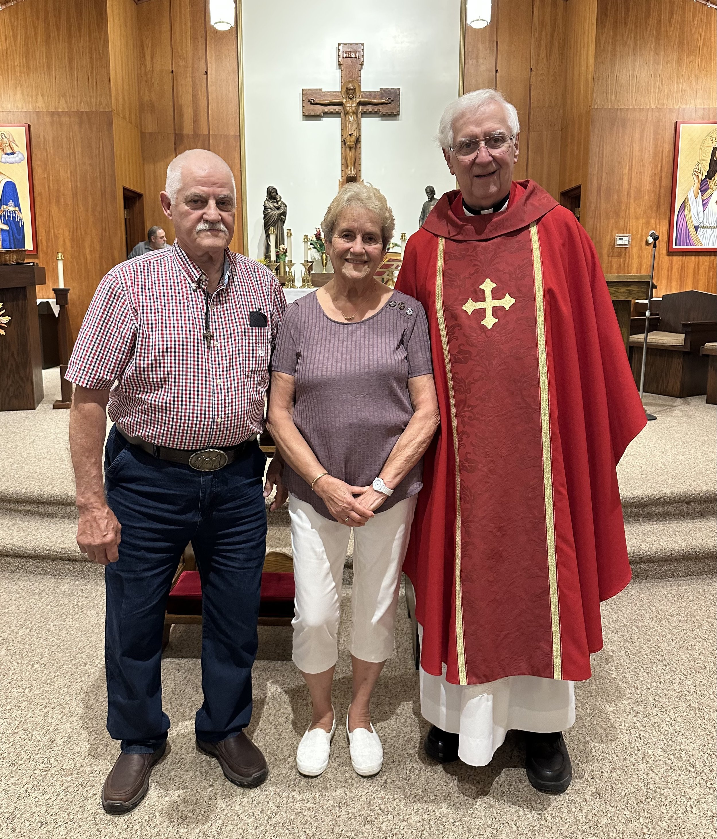 Three people standing inside a church with a crucifix and religious statues. A man in a checkered shirt, a woman in a purple top, and a priest in a red robe are smiling at the camera.