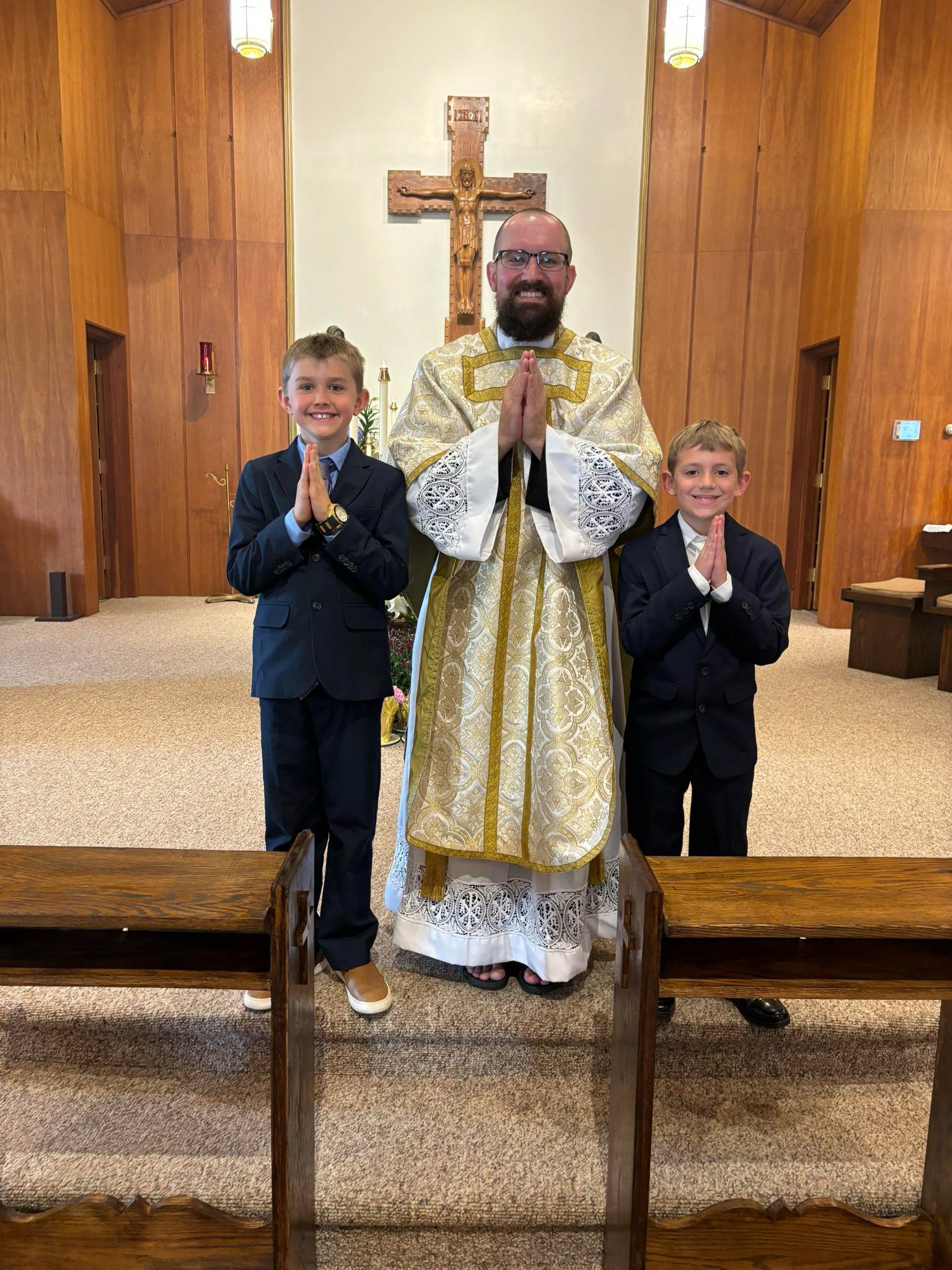 A priest and two young boys in suits are standing in a church, each with hands together in prayer, smiling at the camera. The priest is wearing a gold and white liturgical vestment, and the boys are dressed in dark suits with light collared shirts. T