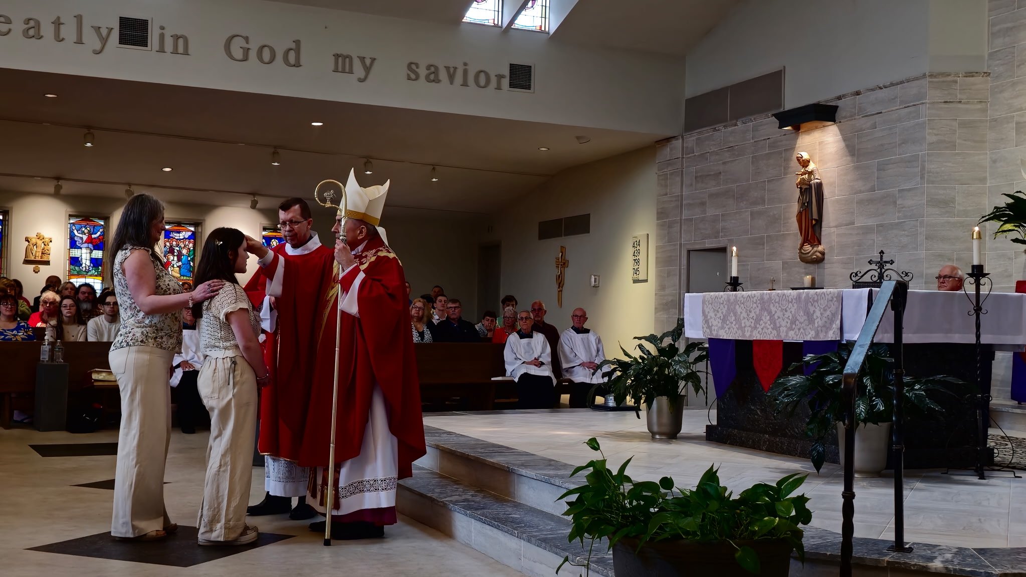 A religious ceremony in a church, where a bishop in red robes is blessing a young girl and woman, with a congregation and altar in the background.