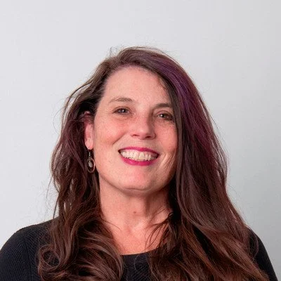 Portrait of a smiling woman with long wavy brown hair and purple highlights, wearing a dark top, against a plain gray background.