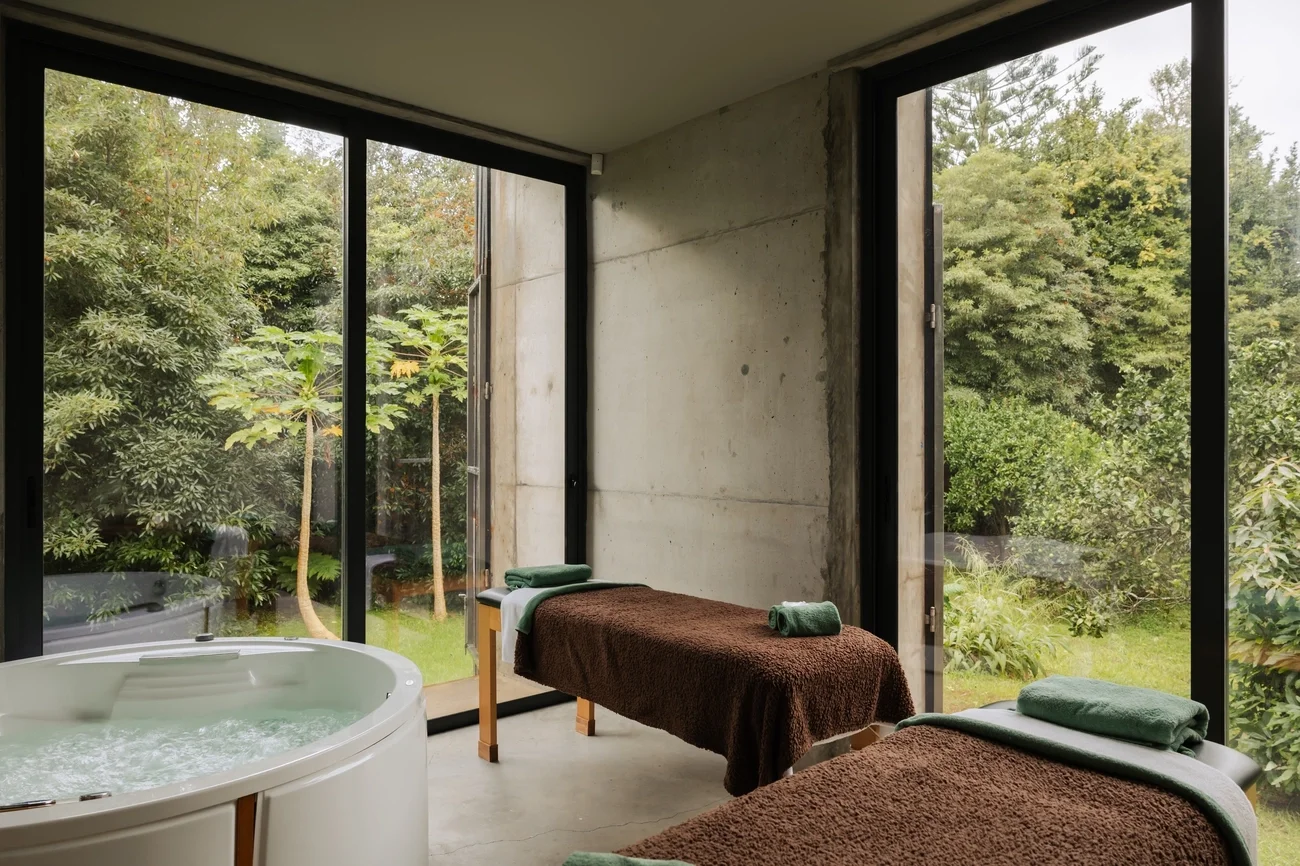 Spa room with two massage tables covered in brown towels, each with folded green towels, and a white hot tub near large floor-to-ceiling windows overlooking a green garden.