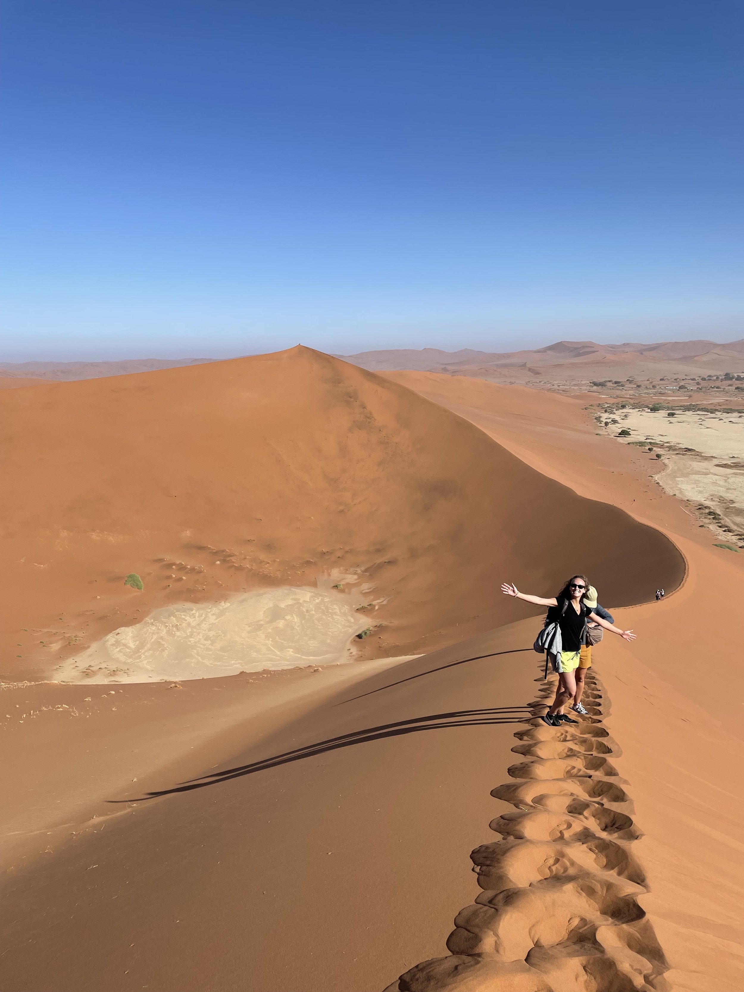 Two women walking with backpacks along a narrow trail in the sand dunes of the Kalahari Desert, with a vast landscape of sand dunes and mountains under a clear blue sky.