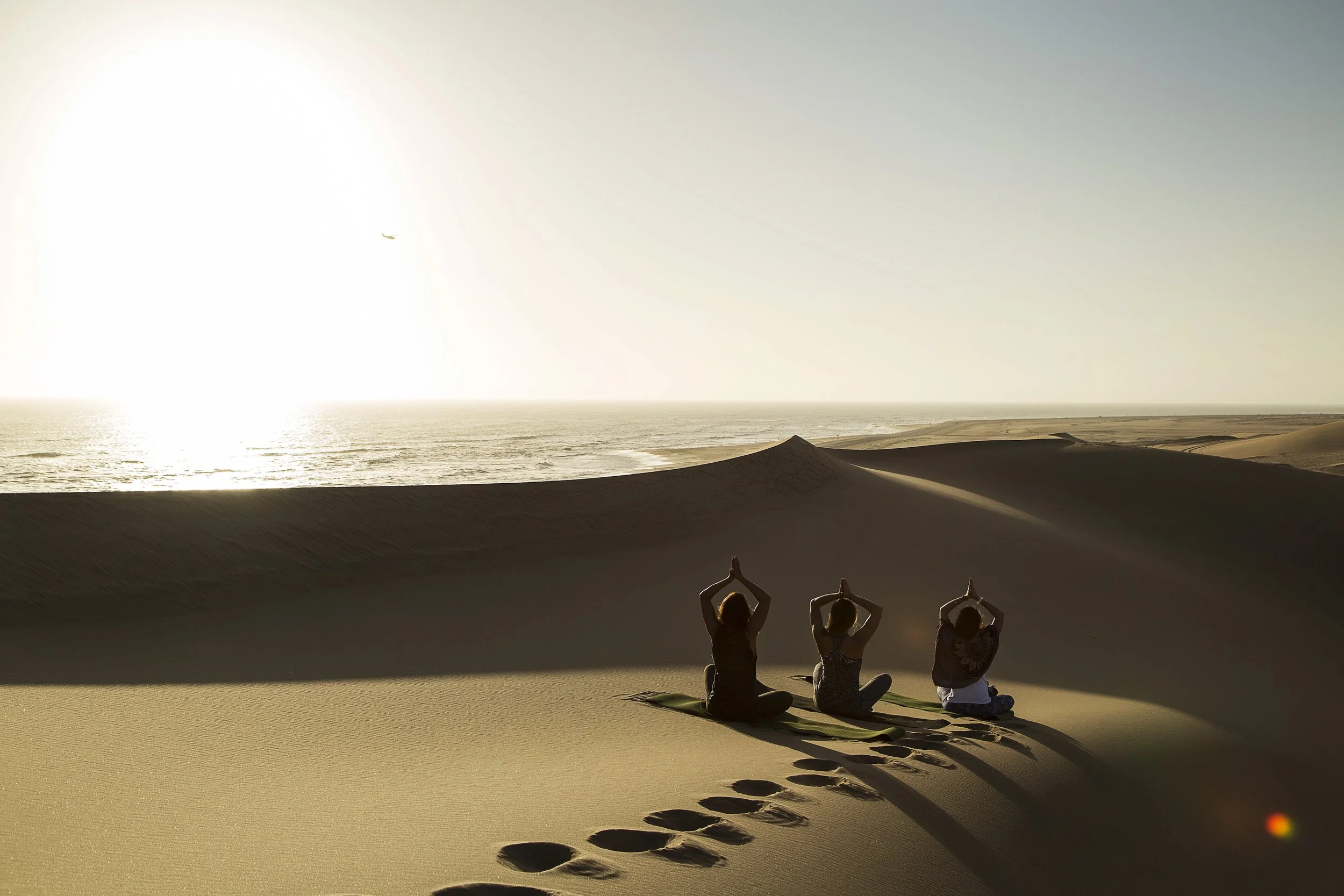 Three people practicing yoga on mats in a desert landscape during sunset, with footprints in the sand leading toward them and an airplane flying in the sky.