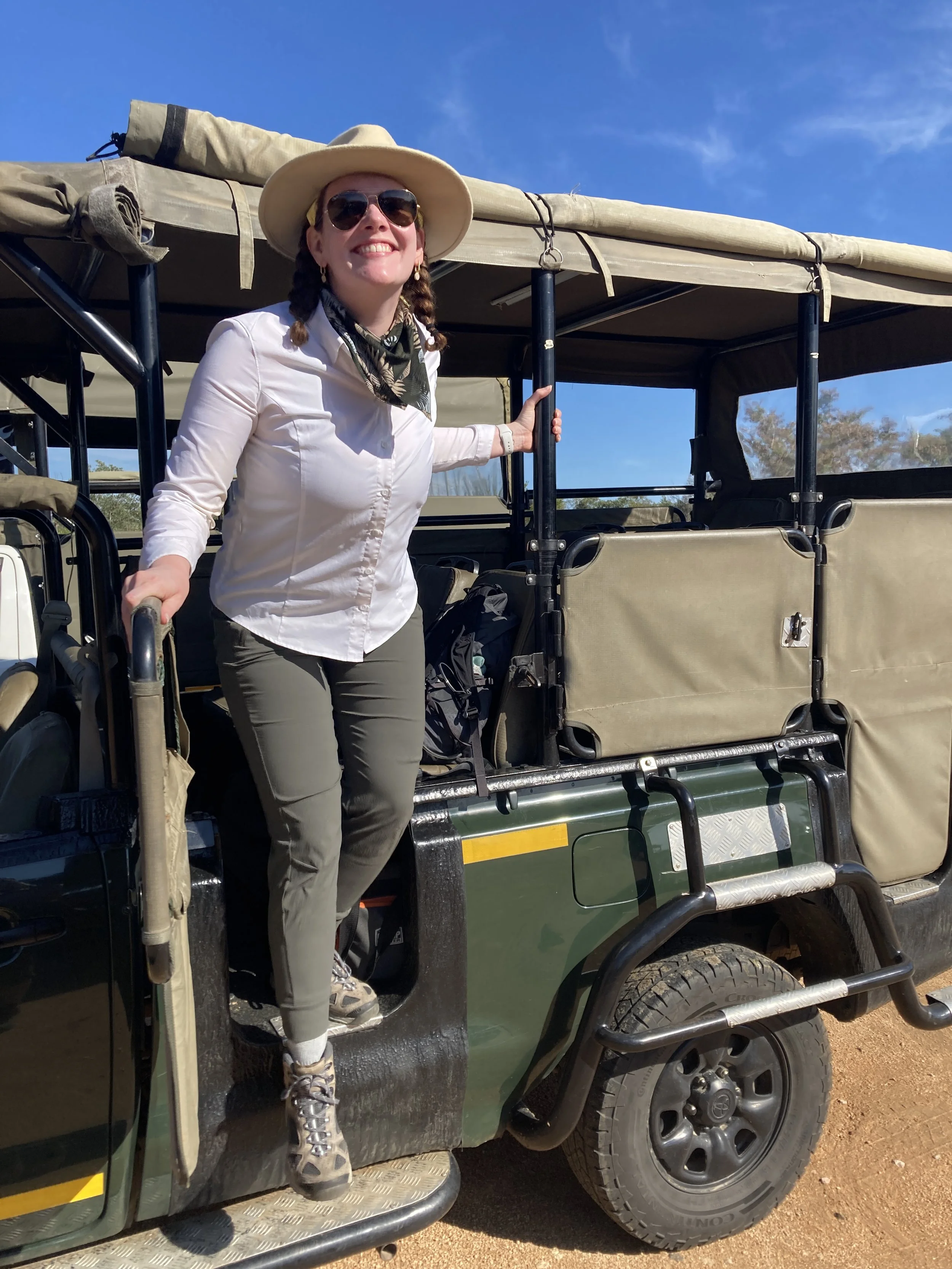 A woman with a wide-brimmed hat, sunglasses, earrings, a white shirt, gray pants, and hiking boots stands on the steps of a safari vehicle, smiling, with her right hand on a black pole and holding a gray handle with her left hand. The vehicle is outdoors, with a blue sky and some trees in the background.