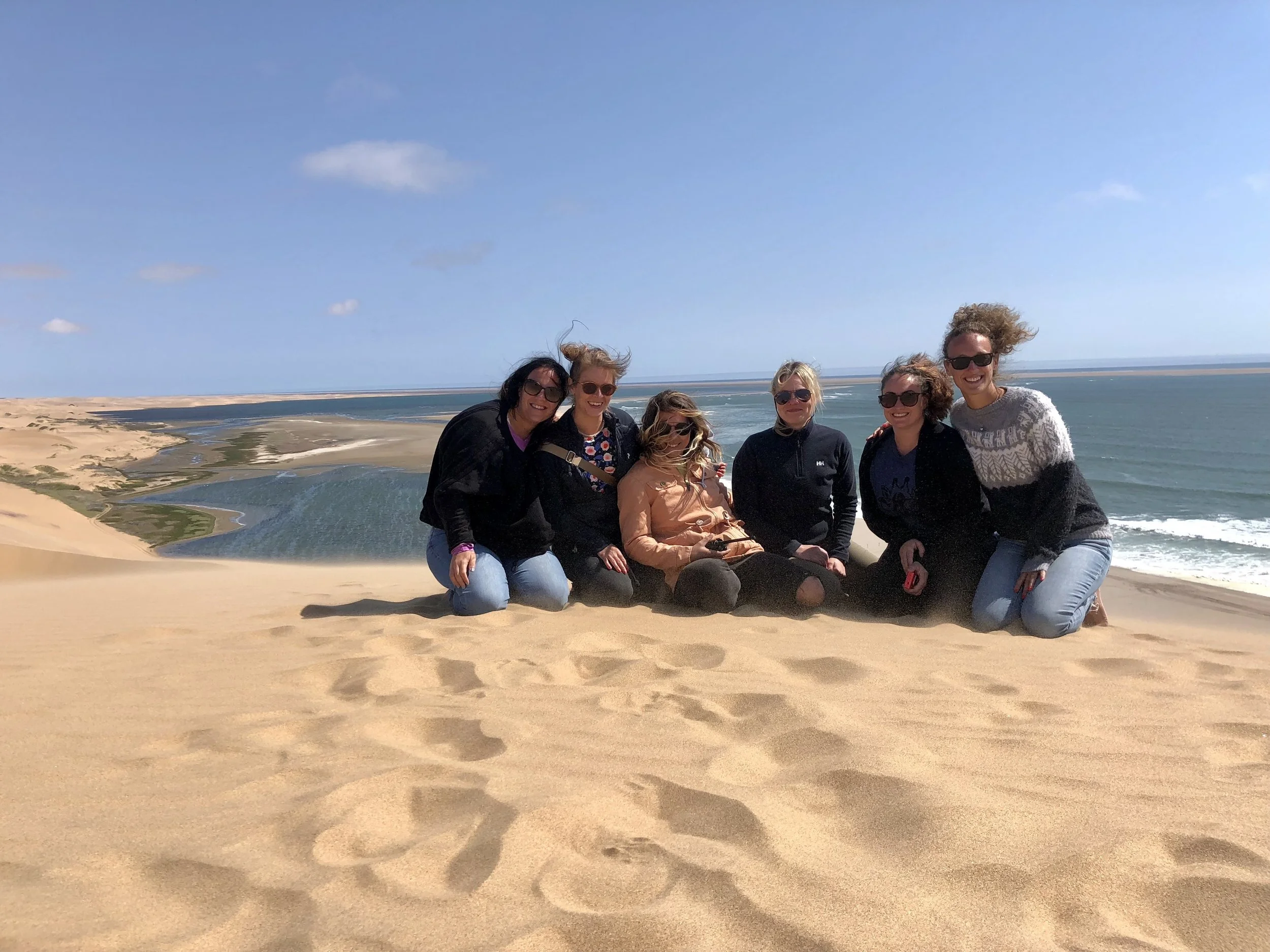 A group of seven women poses on a sandy beach near the ocean in Swakopmund, Namibia, with a clear blue sky overhead and dunes in the background.