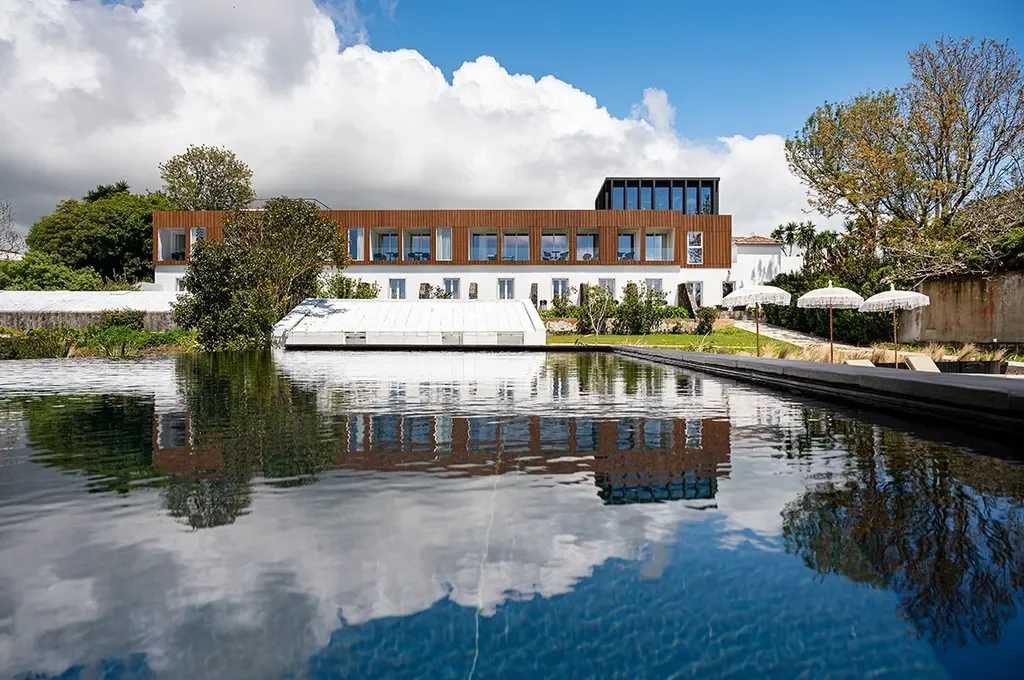 Modern house with a flat roof and large windows, surrounded by trees, with a body of water in the foreground reflecting the house and sky, and umbrellas along the right side.