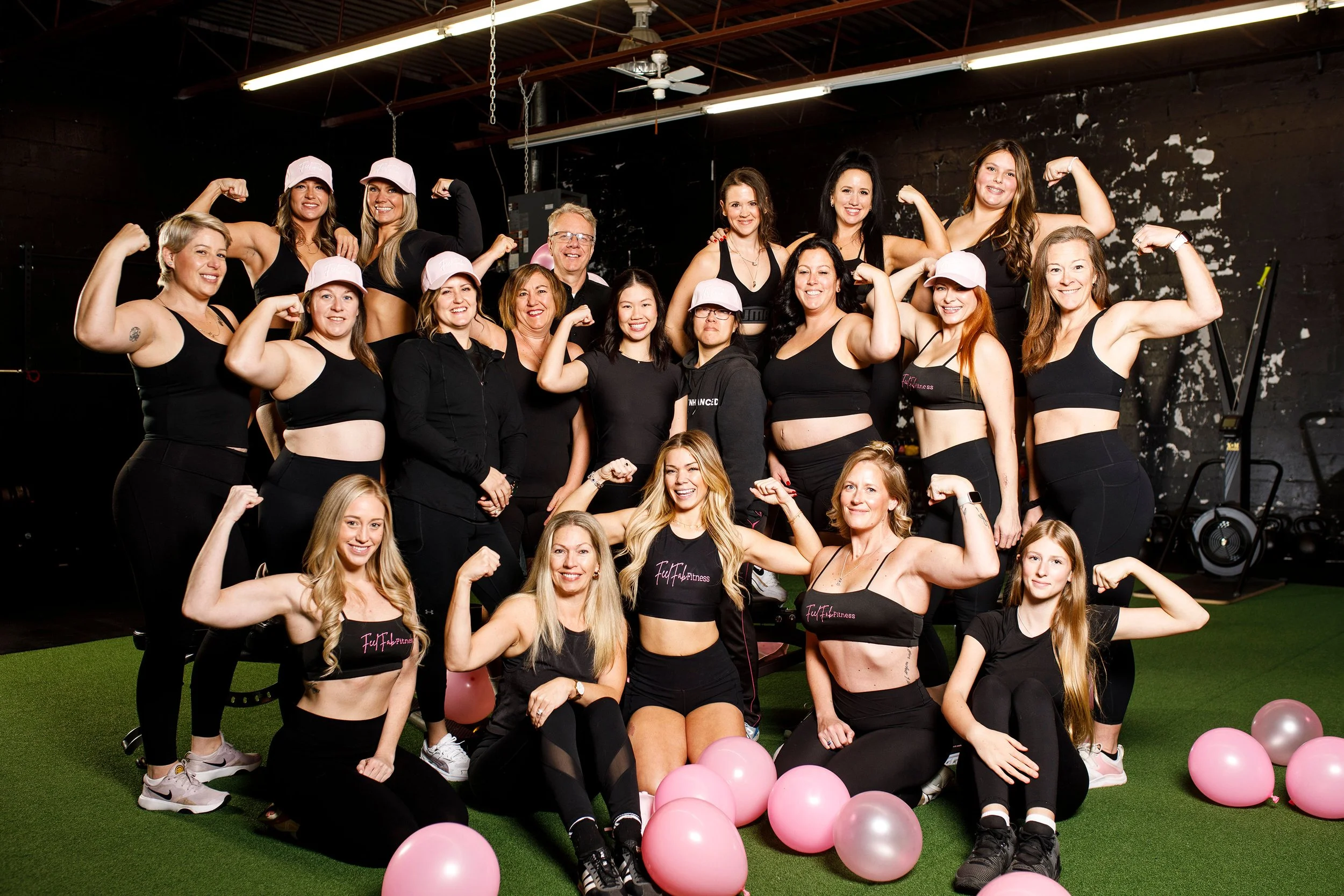 Three women in workout attire, two with black bags that say 'Fulfab Fitness', sitting with their backs to the camera, and one woman facing the camera smiling, kneeling in front of a pink banner that says 'Fulfab Fitness personal training'.