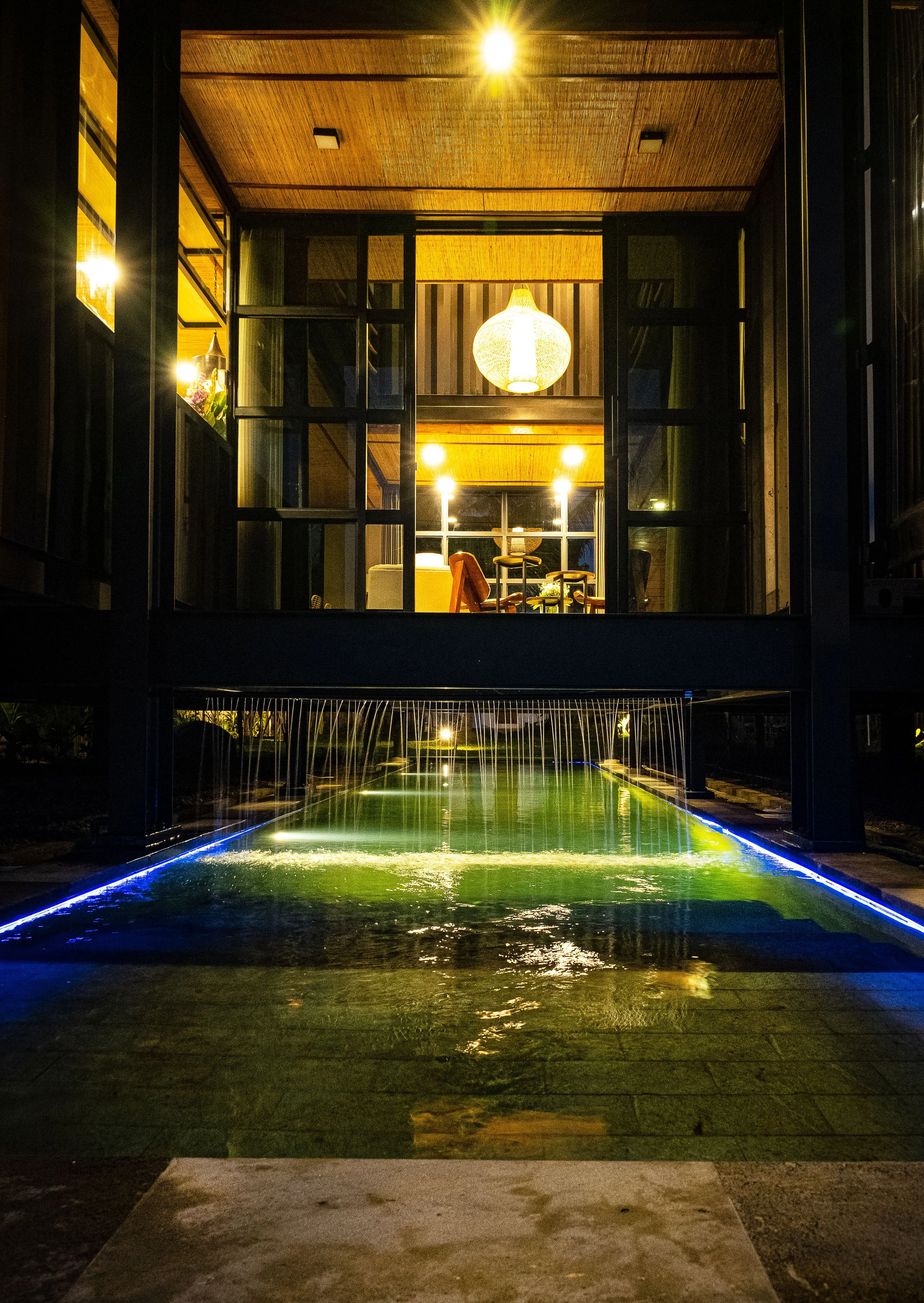 Night view of a modern house with a water feature directly in front, illuminated by blue and white lights, visible through large glass windows that reveal a cozy interior with warm lighting and a hanging lantern.