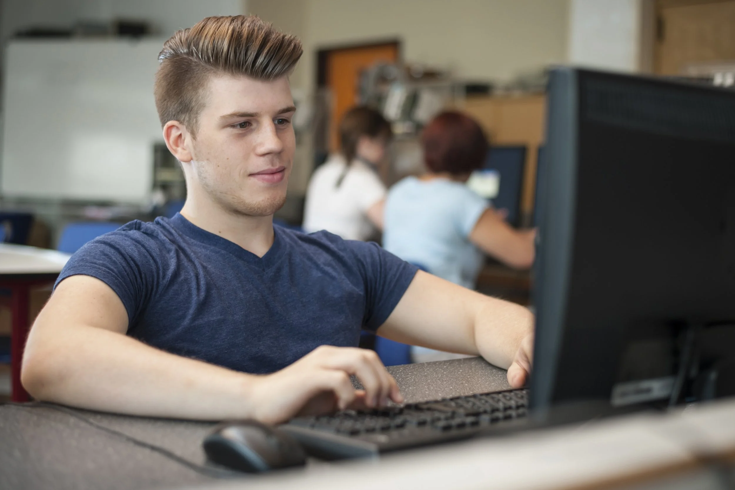 A young man with styled hair wearing a navy blue t-shirt using a computer in a classroom or computer lab with other students working in the background.