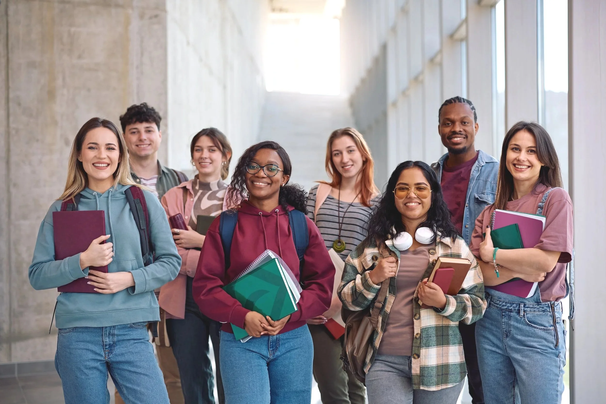 Group of diverse students standing in a school corridor, smiling and holding notebooks and backpacks.