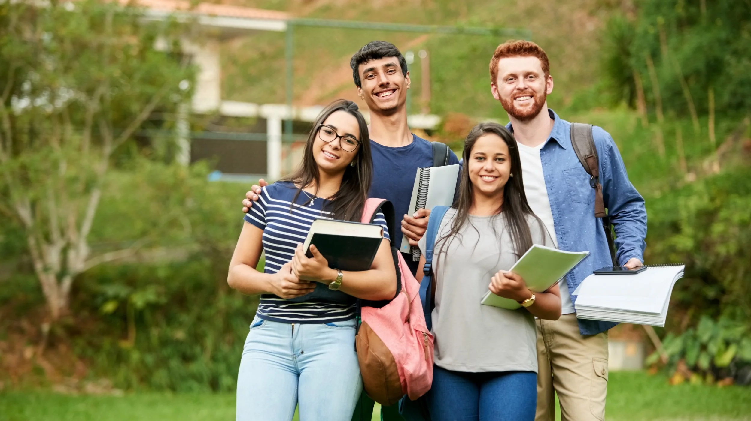 Four college students walking outdoors, smiling, holding notebooks and folders, with greenery in the background.