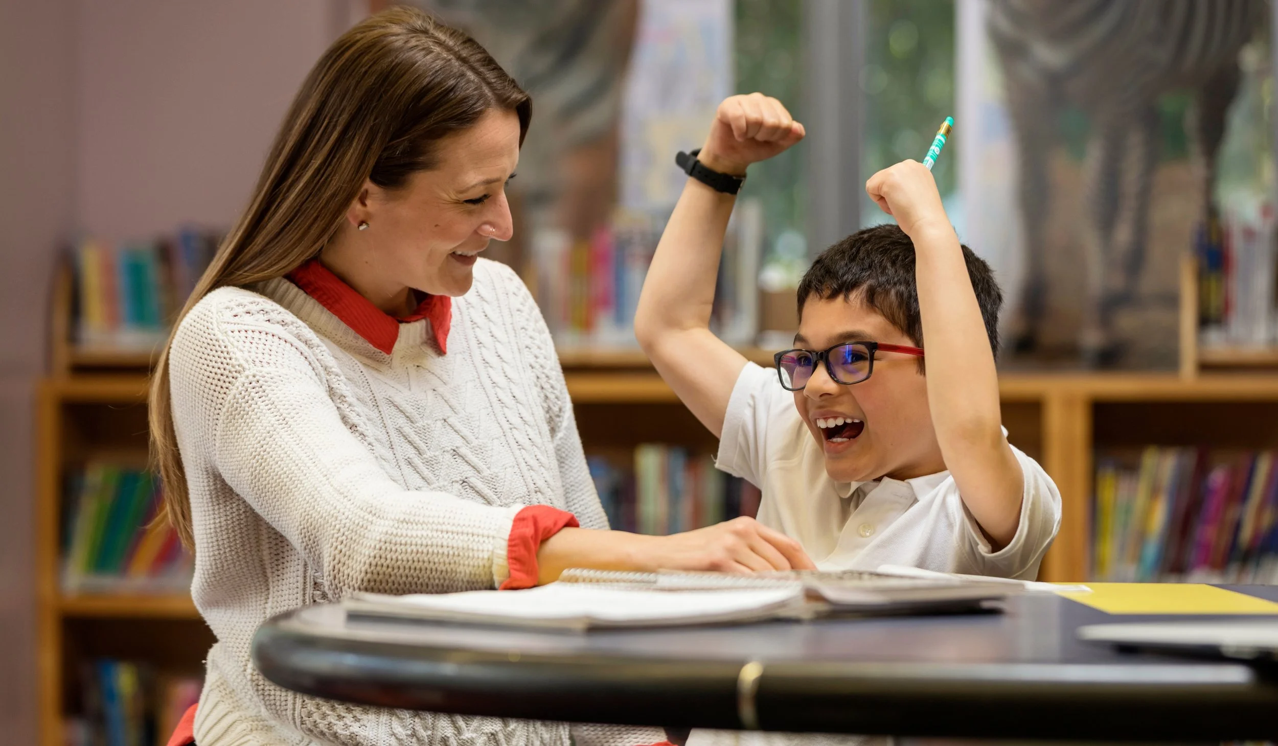 A woman and a boy in a library, smiling and celebrating. The boy is raising his arm with a pencil in hand, showing excitement, while the woman looks on happily.