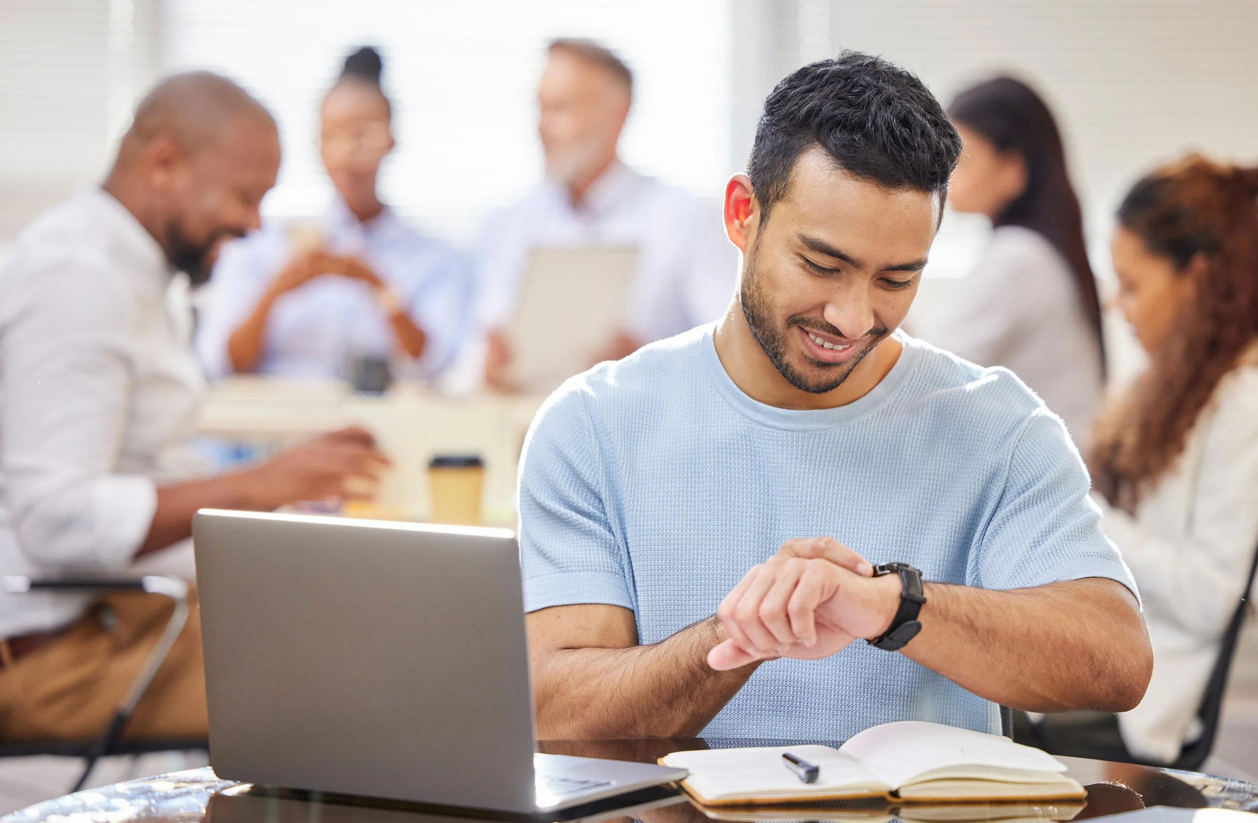 A young man in a light blue shirt looking at his watch and smiling while sitting at a table with an open notebook and a laptop in front of him. In the background, a group of diverse people are engaged in a discussion or working in a bright office setting.
