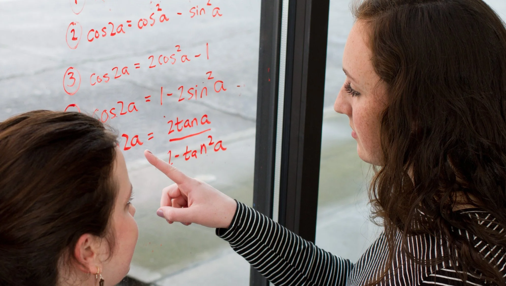 Two women look at a glass window with red mathematical equations written on it. One woman points at the equations, which involve trigonometric functions, while the other listens. Outside, a blurred view of water is visible.