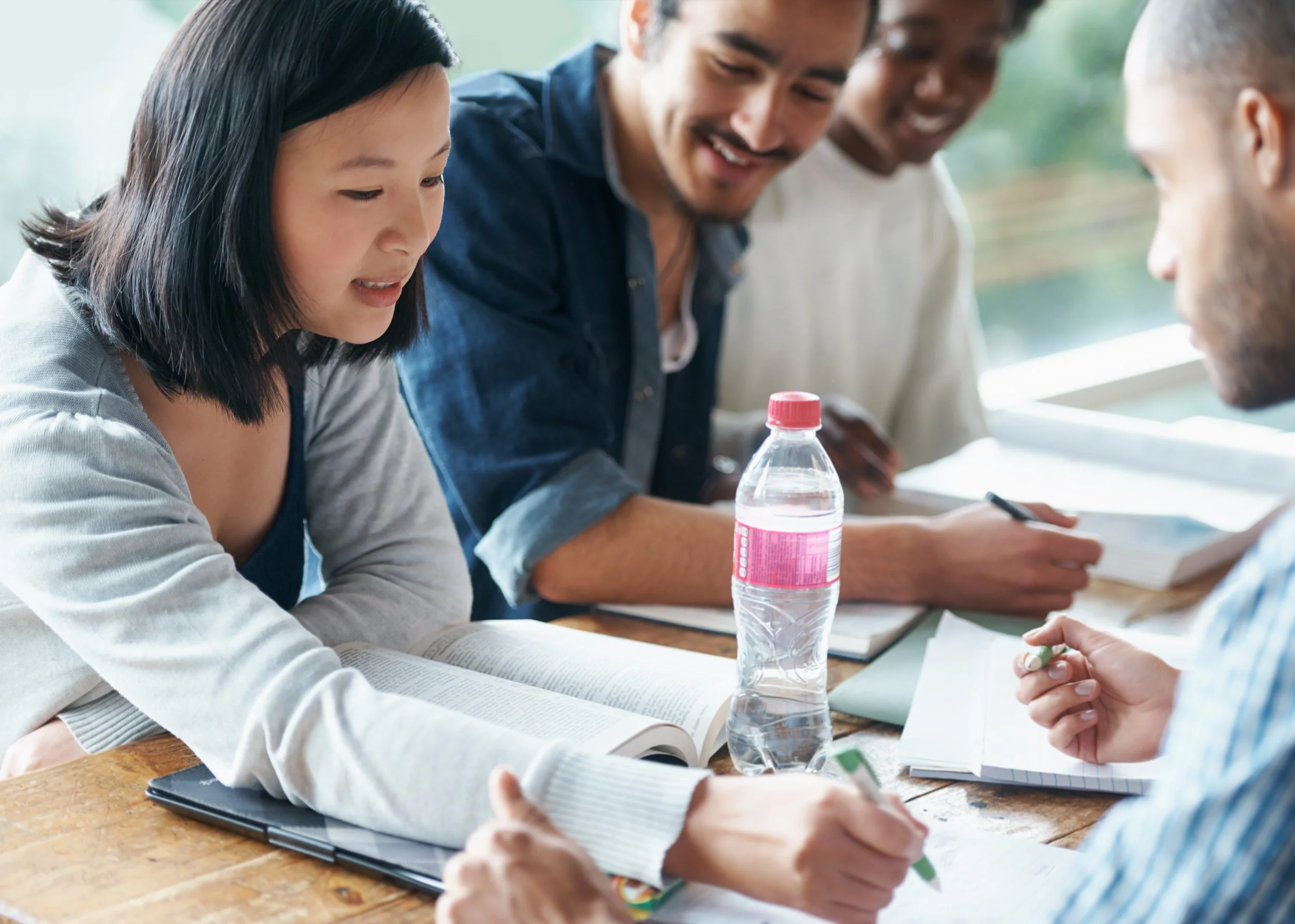 A diverse group of young adults studying and working together at a table with books, notebooks, pens, and a water bottle, smiling and engaged in discussion.