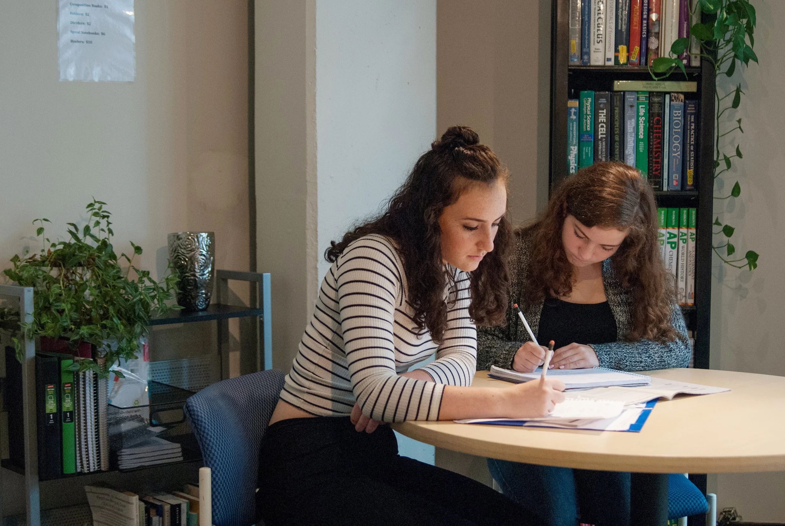 Two young women studying together at a table in a library or study room, with bookshelves and plants in the background.
