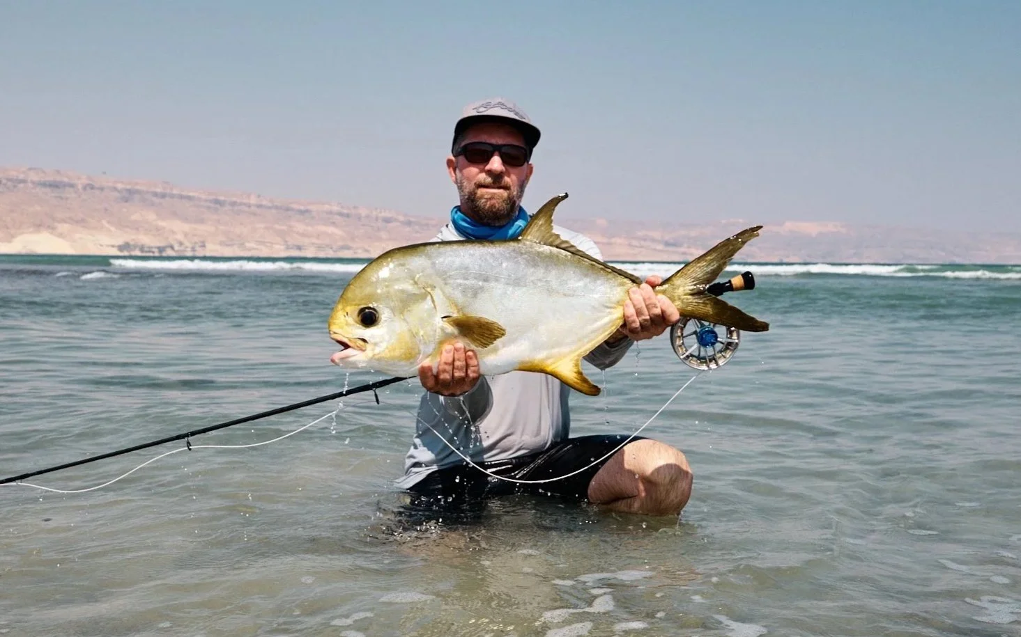A man wearing sunglasses and a hat kneeling in shallow water at the beach, holding a large fish with a silver and yellowish body, a rounded shape, and a prominent eye.