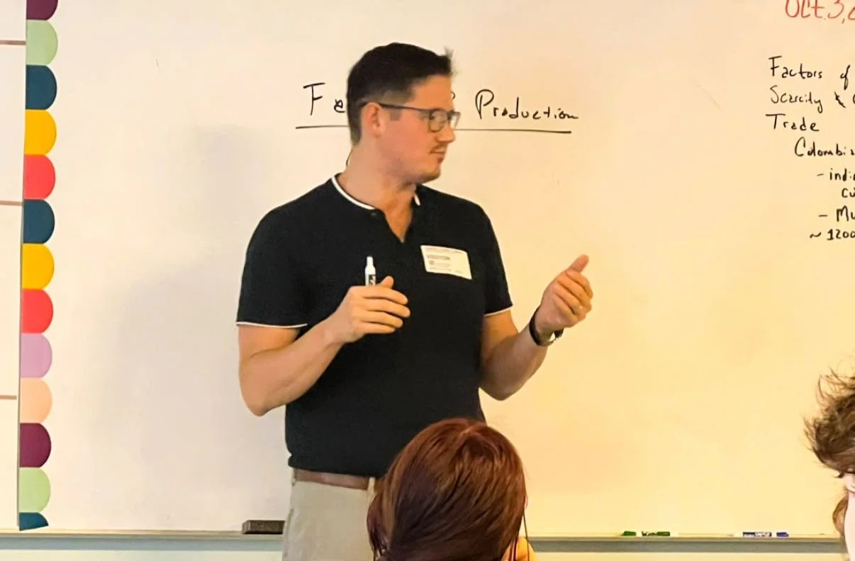 A man with dark hair, glasses, wearing a black polo shirt, standing in front of a whiteboard, giving a presentation.
