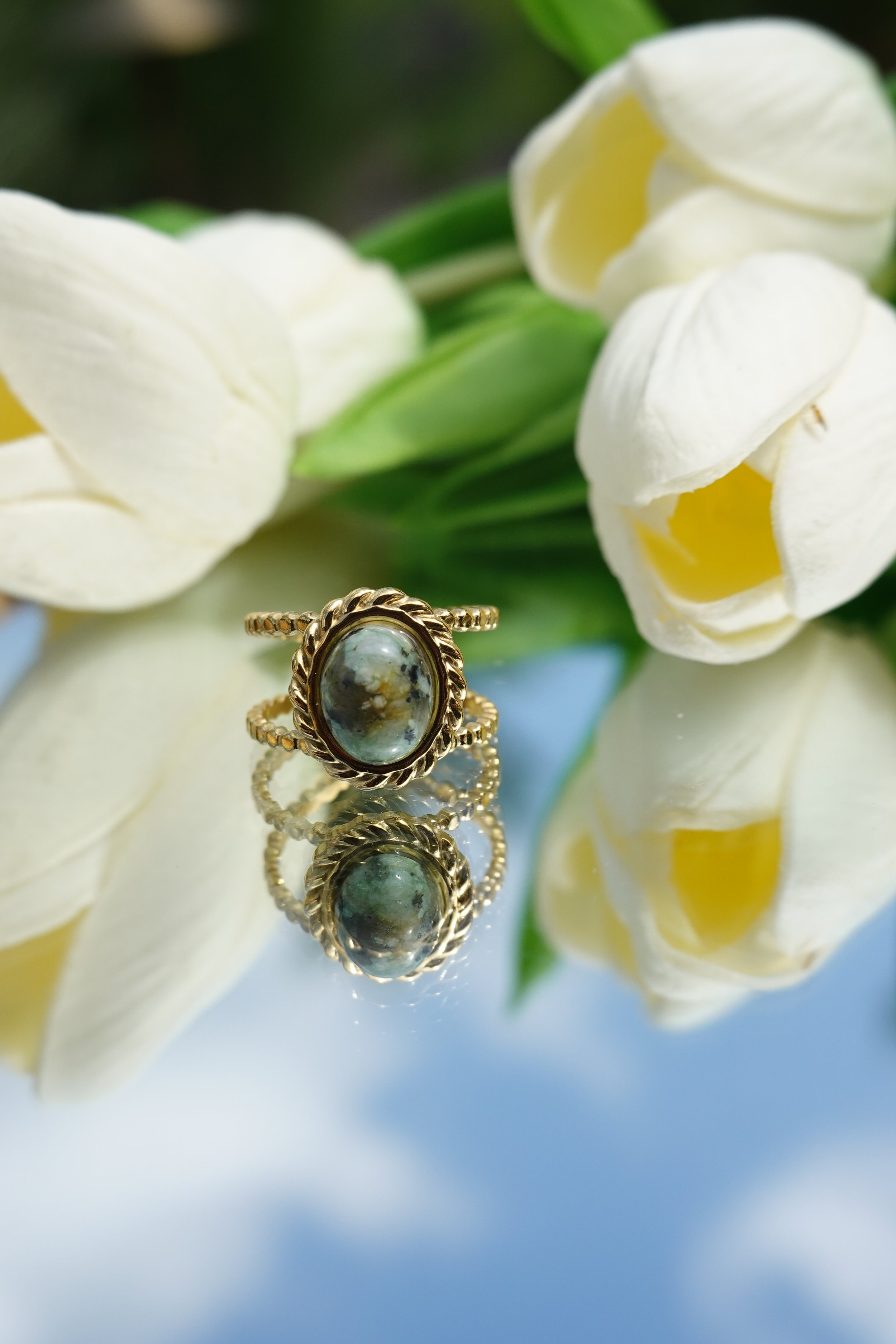Close-up of a gold ring with an oval gemstone, reflected on a shiny surface, surrounded by white flowers in the background.