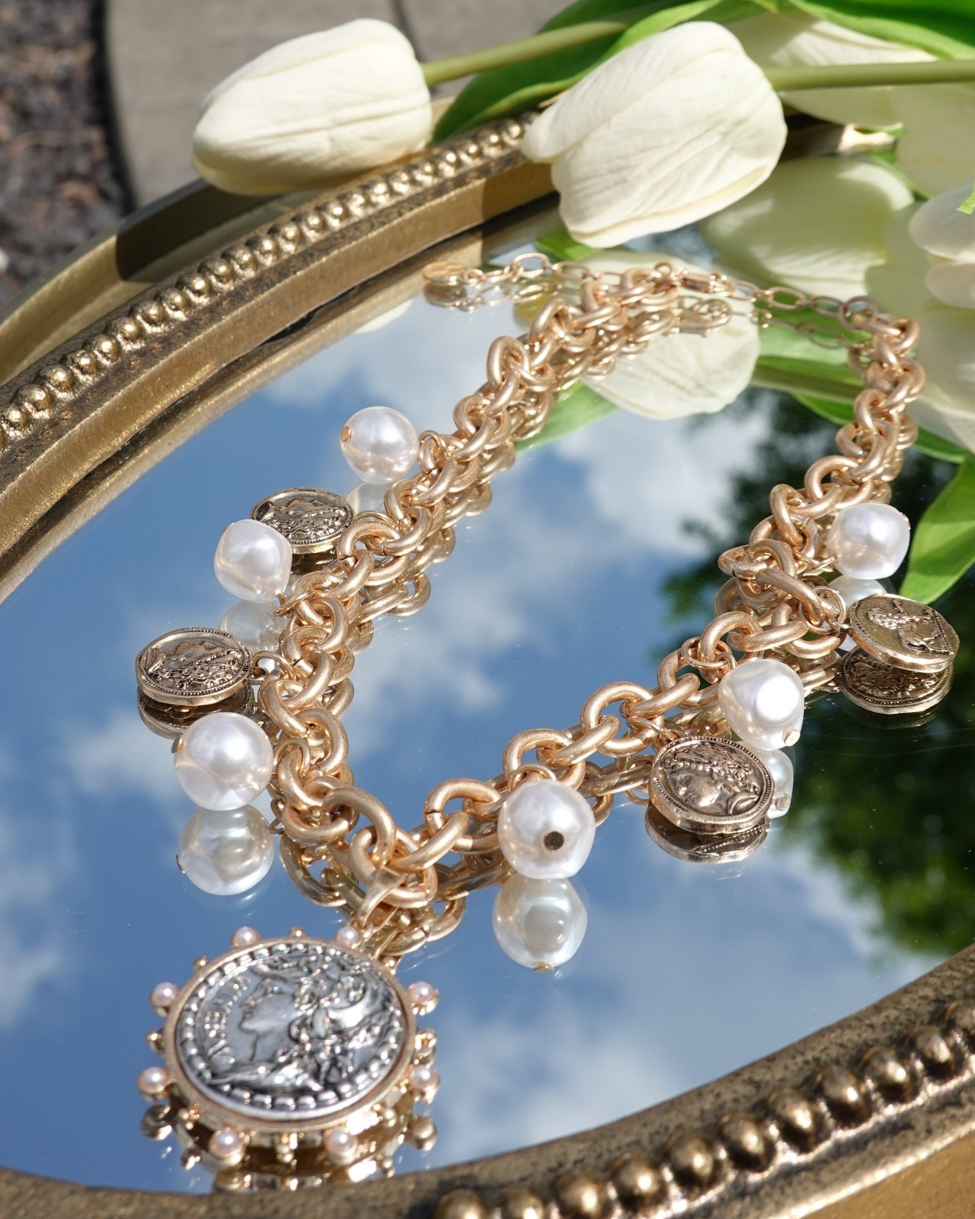 Gold charm bracelet with pearl and coin charms resting on a mirror tray surrounded by white tulips.
