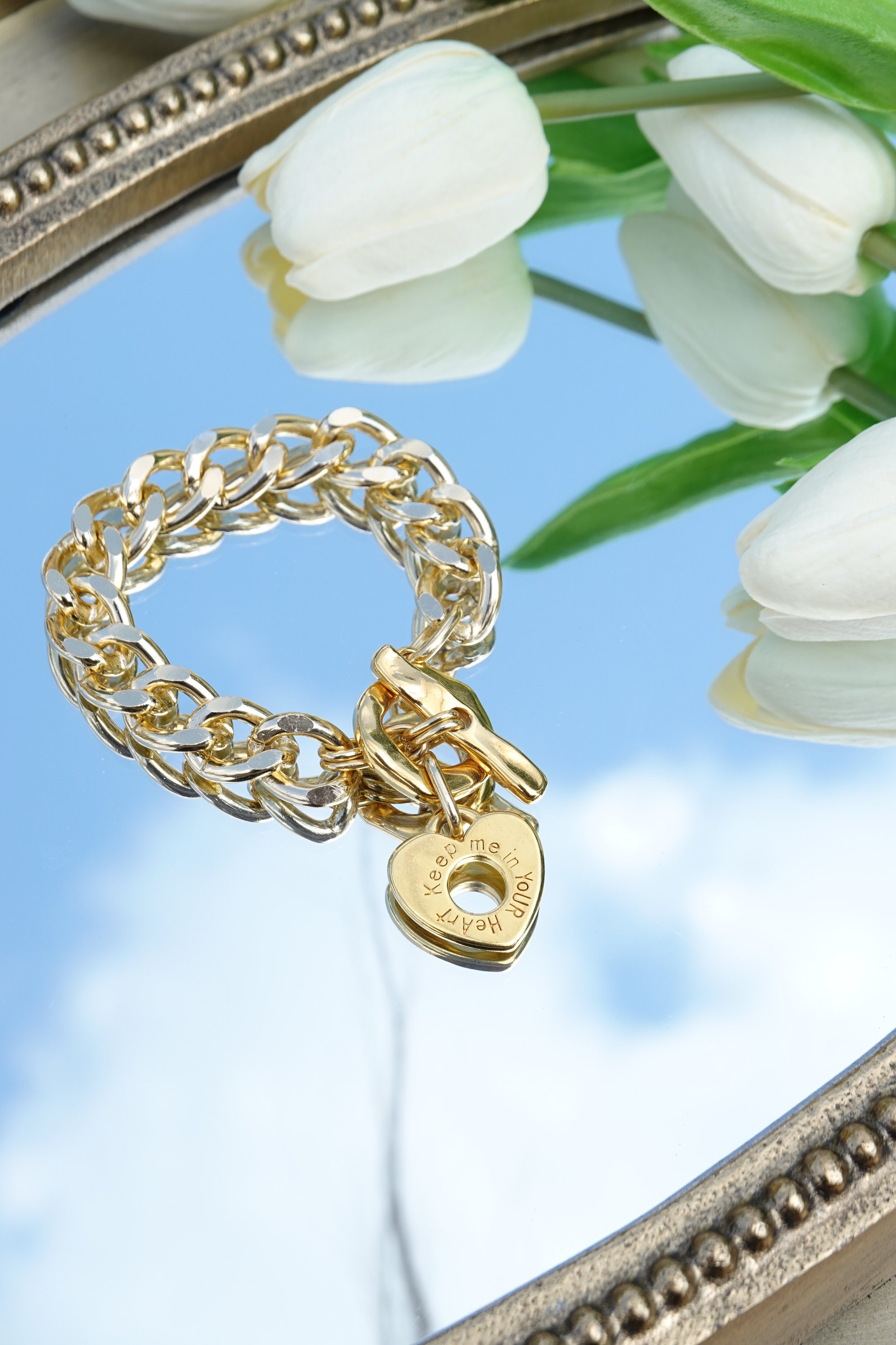 A gold chain bracelet with a heart-shaped tag inscribed "Keep me in your heart" resting on a mirrored surface with white tulips nearby.