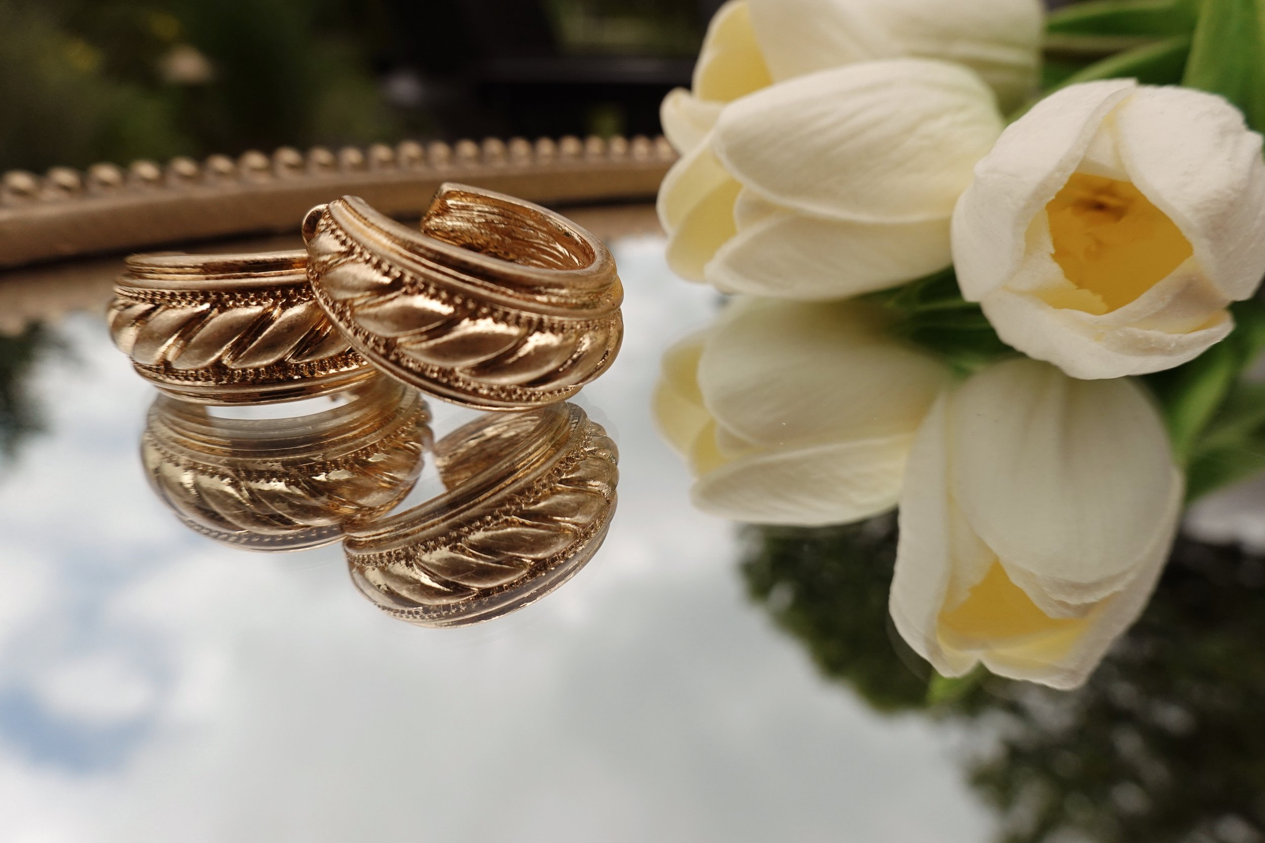Close-up of three rose gold rings with twisted design, reflecting on a mirror surface, with white flowers in the background.