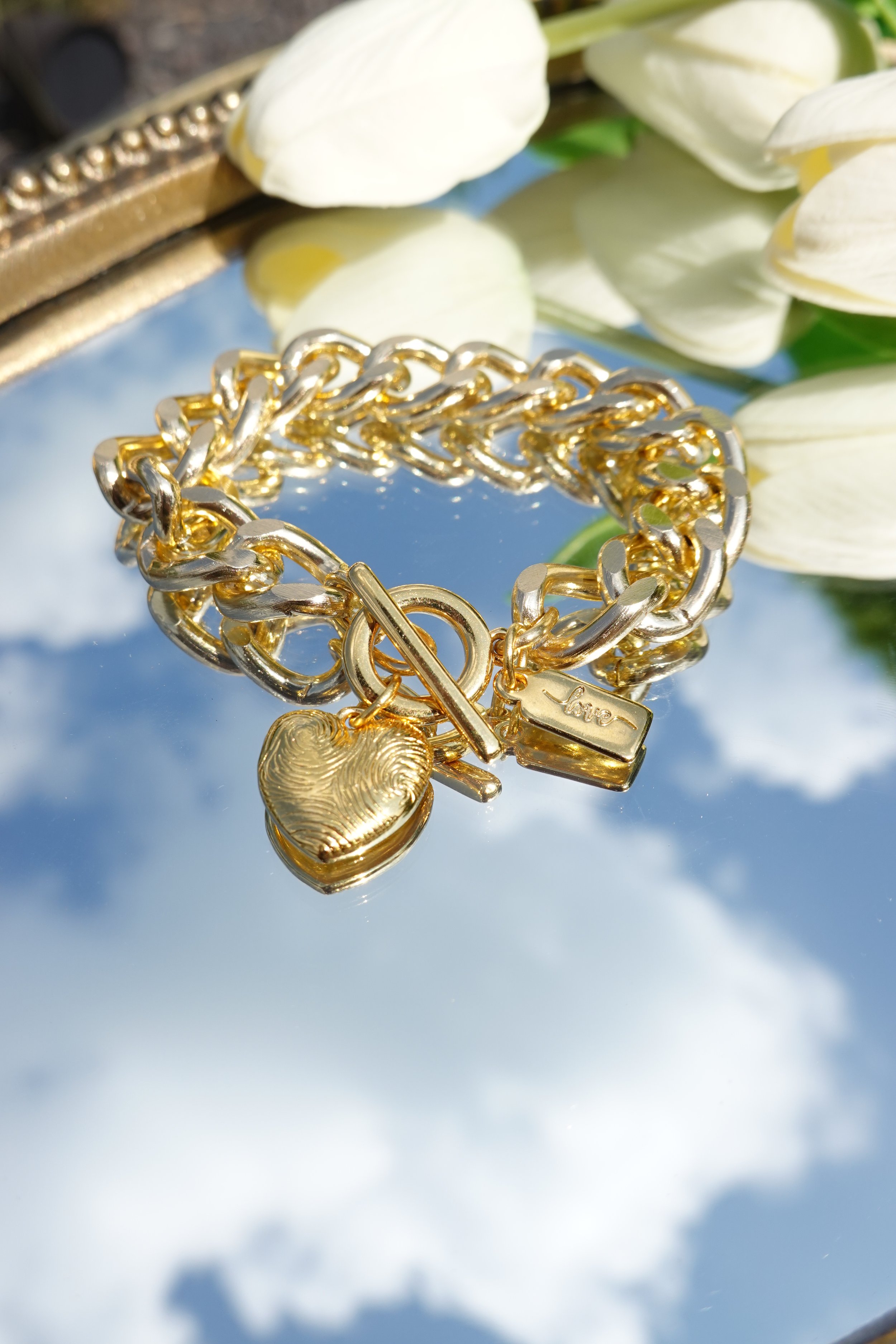 A gold bracelet with charms, including a heart with a fingerprint design, a rectangular tag engraved with "love," and a toggle clasp, resting on a reflective surface with white tulips in the background.