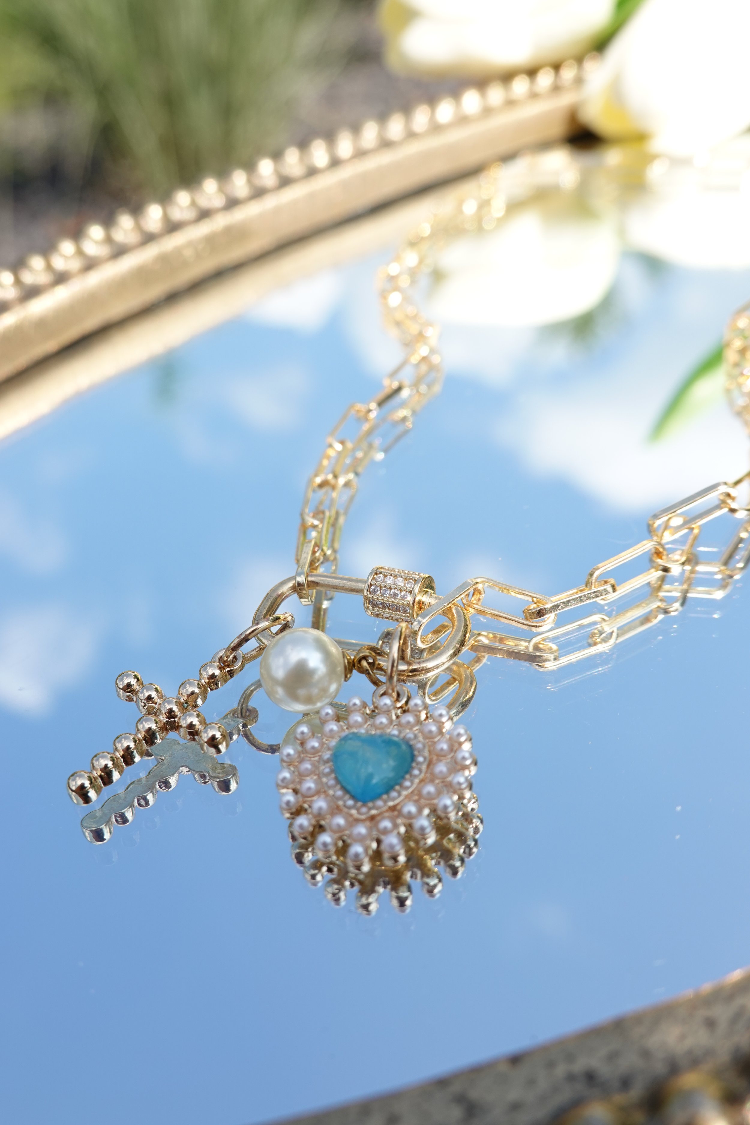 Gold jewelry necklace with charms, including a cross, pearl, and heart-shaped gemstone, resting on a reflective mirrored surface.