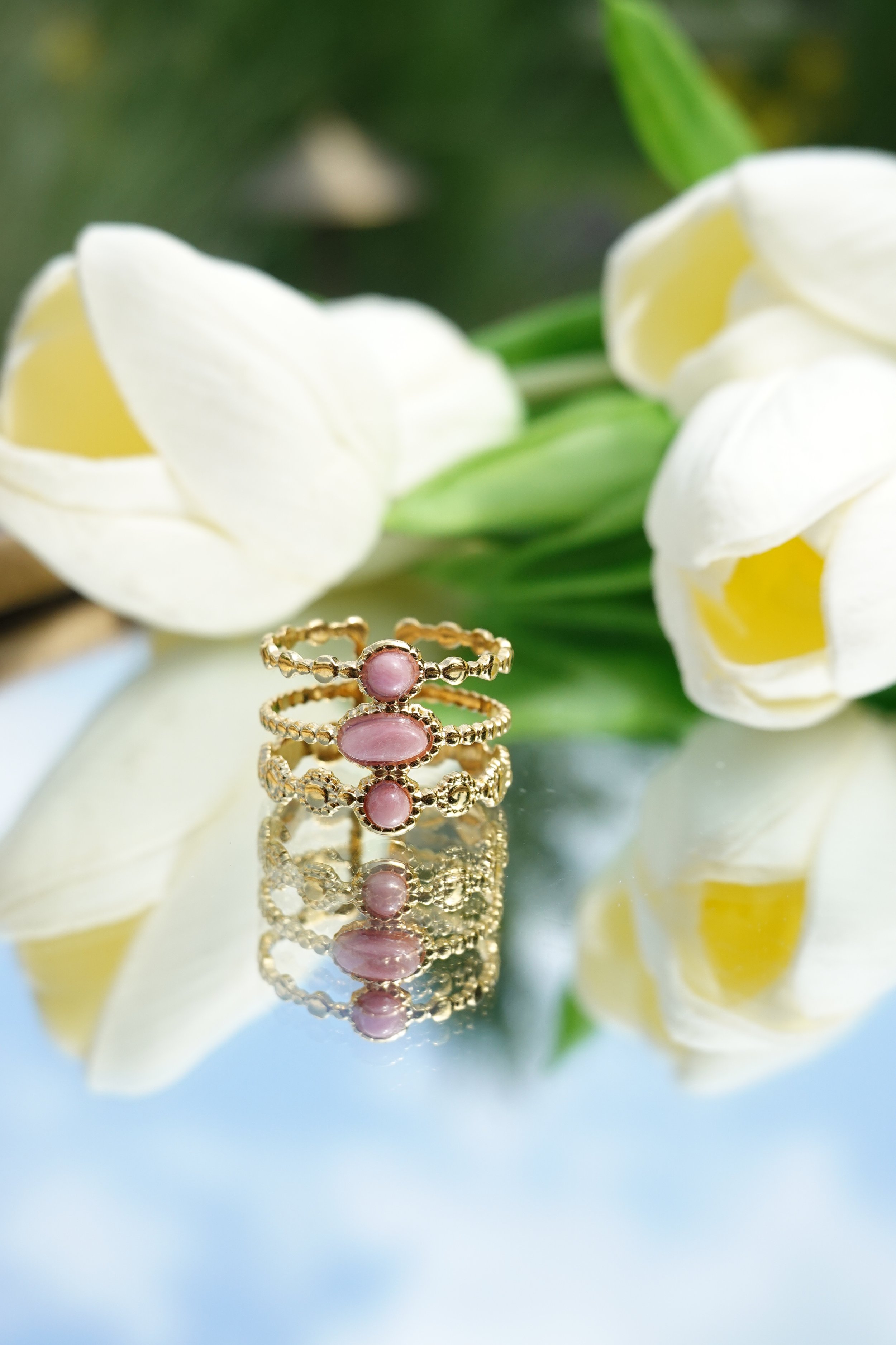 Three gold rings with pink gemstones on a reflective surface, with white and yellow flowers in the background
