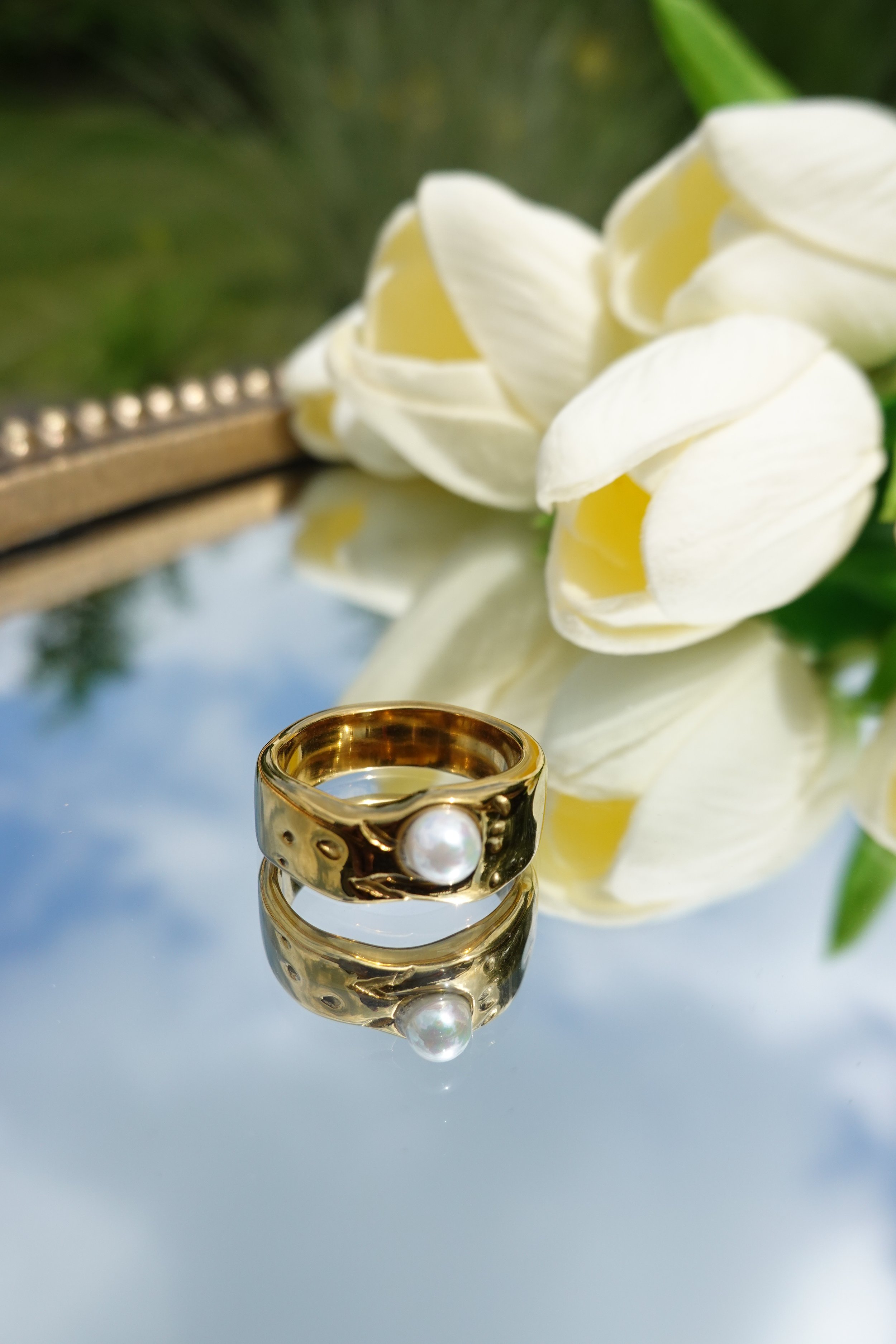 A gold ring with a pearl on a reflective surface, with white tulips and greenery in the background.