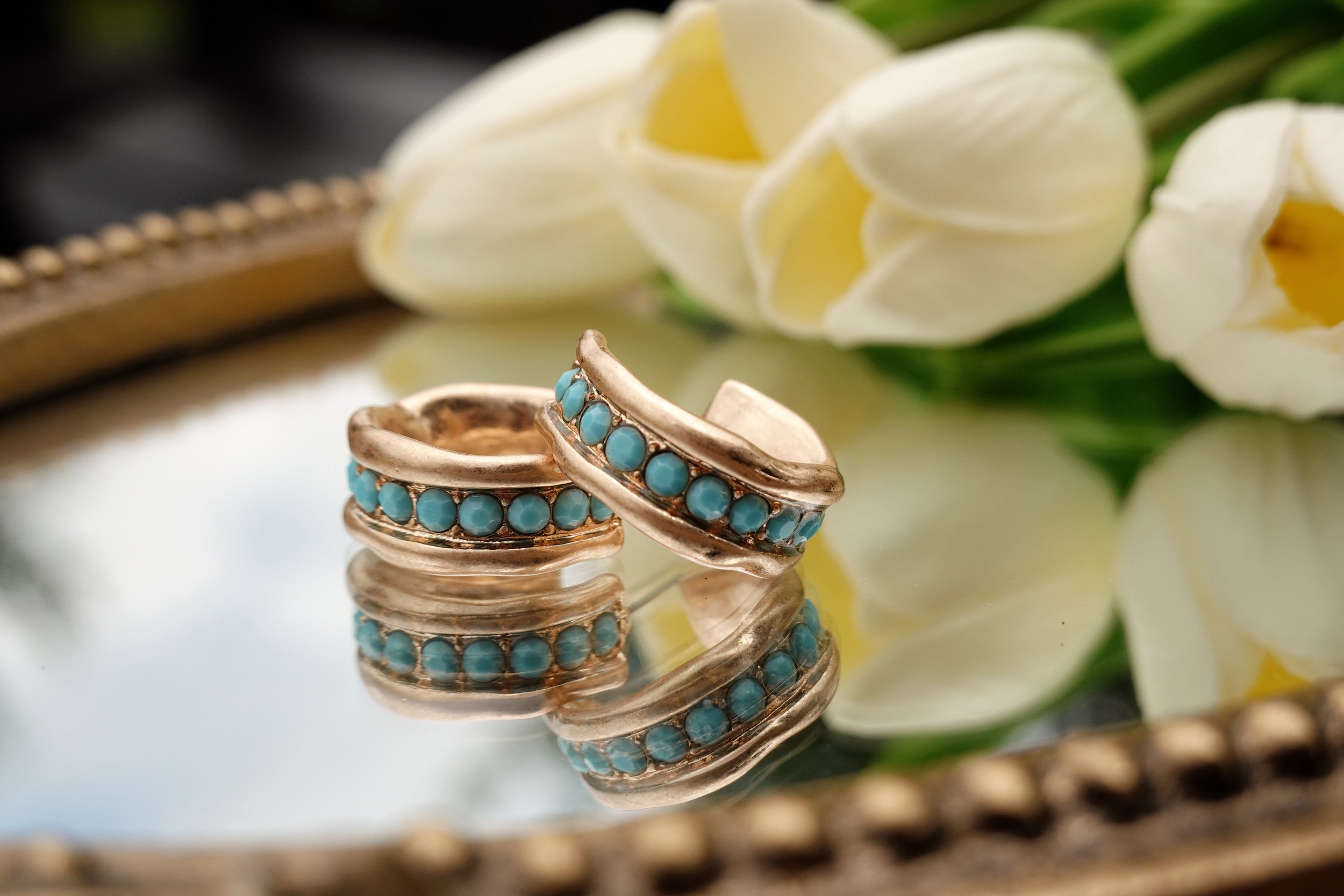Gold ring with blue stones on a reflective surface, with white flowers in the background.