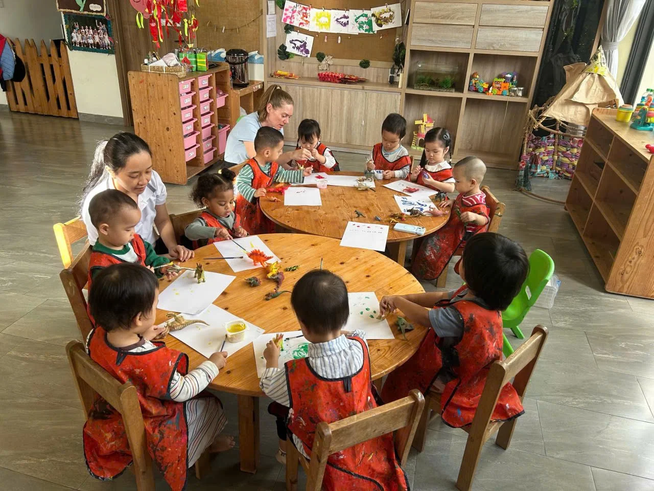 Children and teachers in a classroom making art projects at a round wooden table.