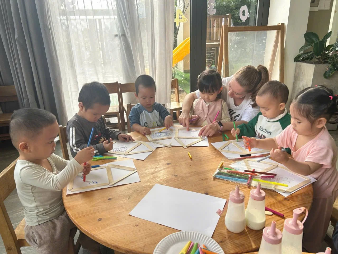 A group of young children and a teacher are seated around a circular wooden table, engaging in arts and crafts activities involving colorful markers and paper. The setting appears to be a classroom or daycare, with natural light coming through large 