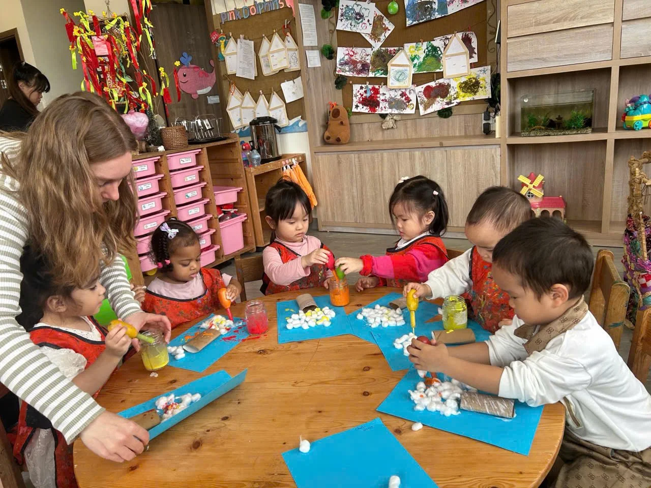 Children participating in a crafting activity at a wooden table, supervised by an adult, with cotton, colorful paints, and paper on the table. Classroom decorated with children's artwork and colorful decorations in the background.