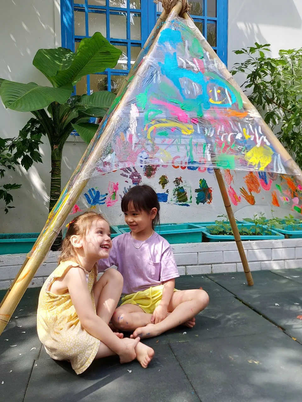 Two young girls sitting on the ground smiling and talking under a homemade transparent tent with colorful paint strokes. A large green plant and a white wall with blue window panes are in the background.