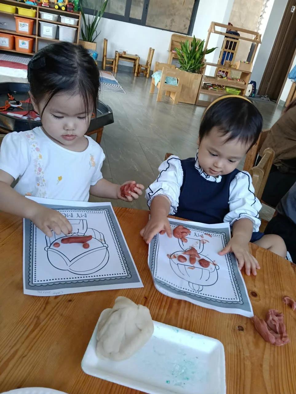 Two young children sitting at a wooden table engaged in a play activity with cookie dough and printed coloring pages. The girl on the left has dark hair tied back, wearing a white shirt, and the boy on the right has dark hair, wearing a navy blue and