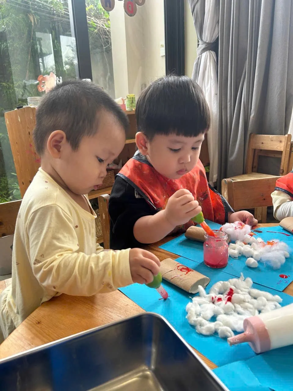 Two young children sitting at a wooden table, engaging in a craft activity with cotton, paint, and paper. They are focused and working with their art supplies.
