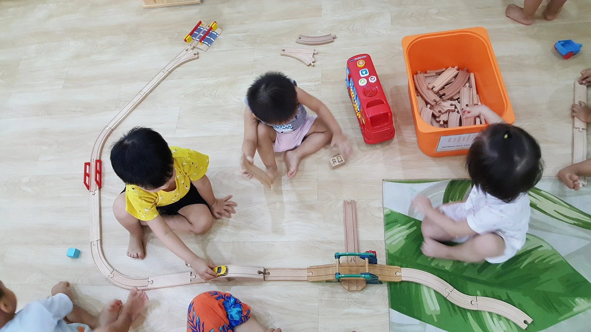 Children playing with a wooden train set on a light-colored floor, with a green mat, orange storage bin, and various train track pieces around them.