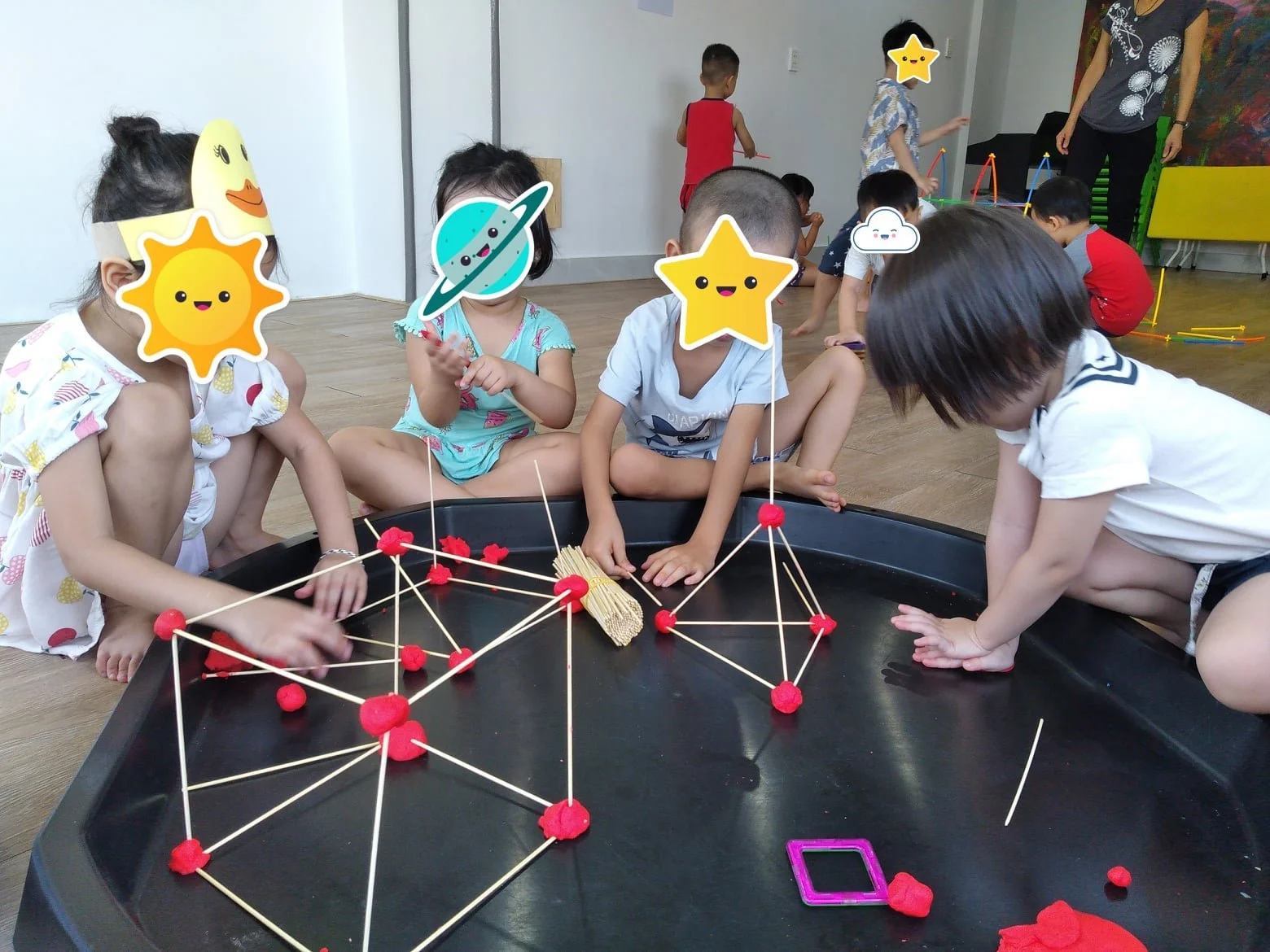Children playing with red yarn and toothpicks to build geometric structures on a black tray, some children have cartoon star, sun, planet, and cloud stickers on their faces, in a classroom setting.