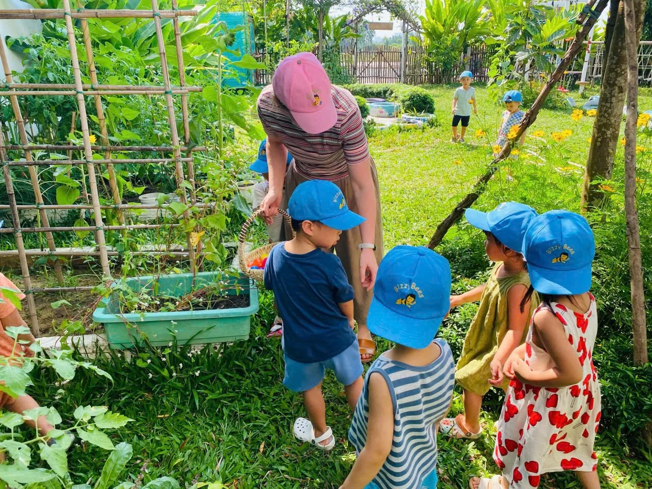 A woman in a pink cap shows children in blue hats how to care for plants in a garden.