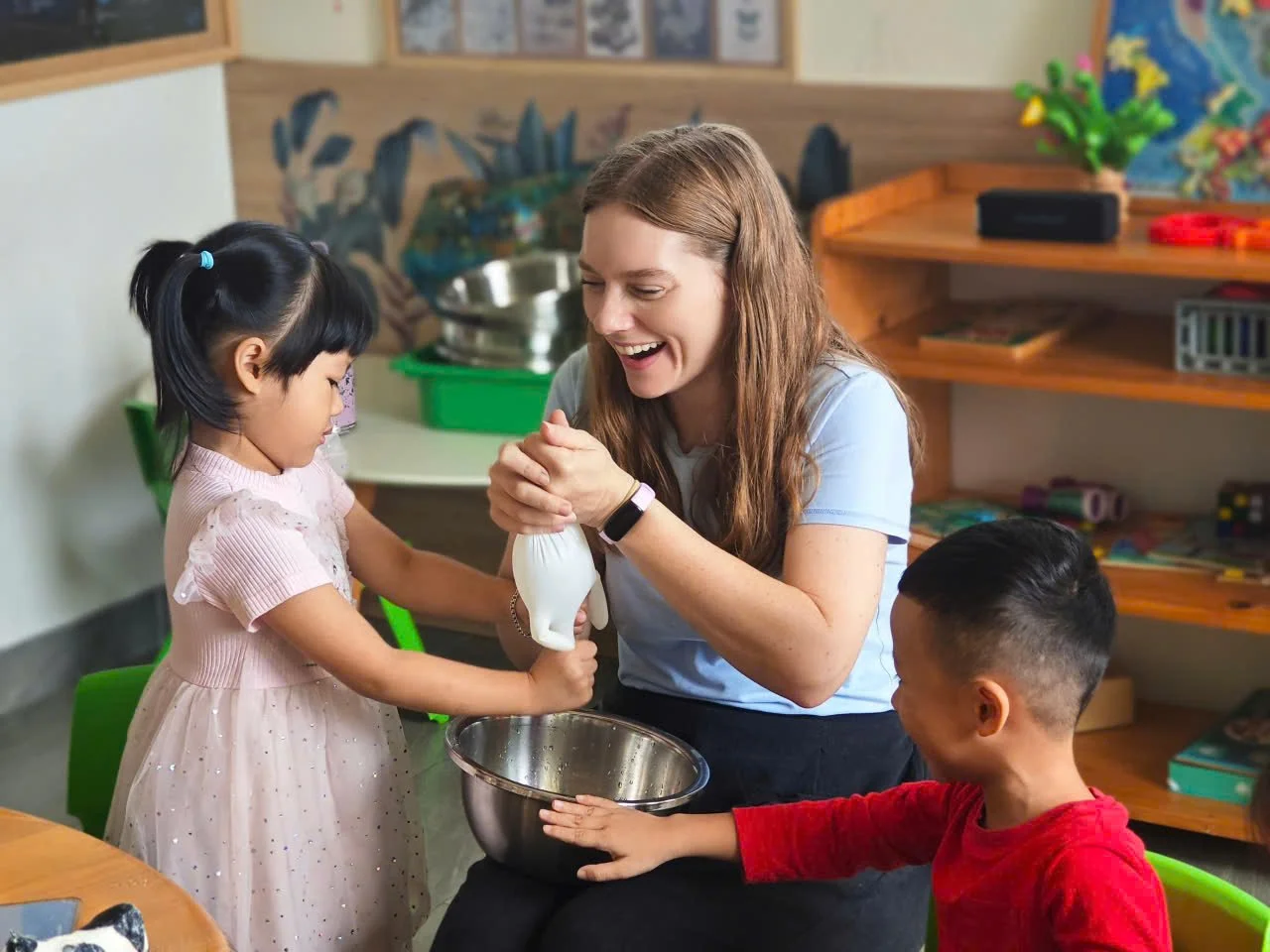 A young woman helping a girl and boy with an activity in a classroom setting.